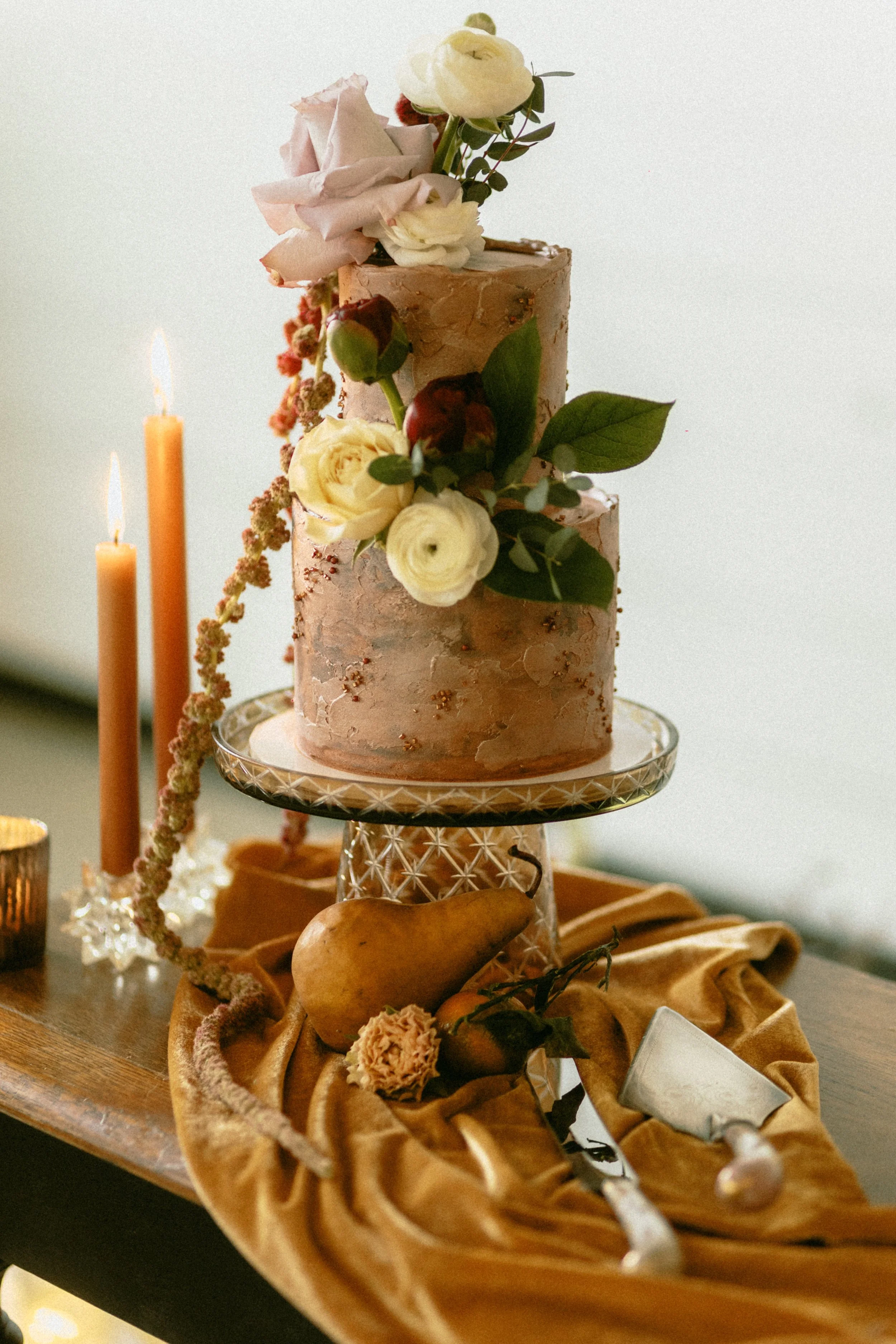 A two-tiered pink wedding cake decorated with flowers and greenery, displayed on a glass cake stand, surrounded by candles, pears, a flower, a cake knife, and a small dish on a draped fabric.