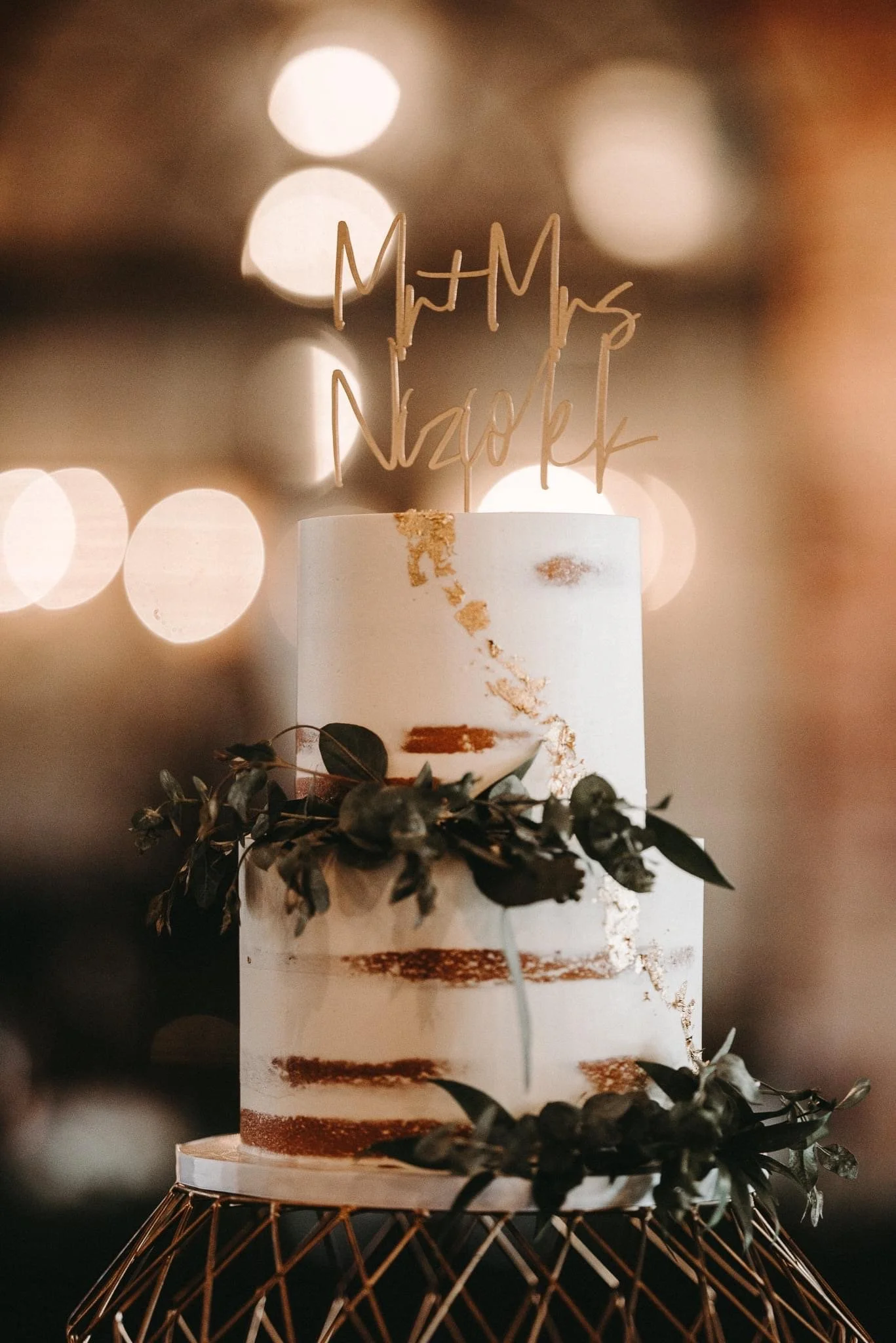 A three-tiered wedding cake with white and gold decoration, topped with a gold 'Mr & Mrs Nazock' cake topper, decorated with green foliage, on a decorative gold stand.