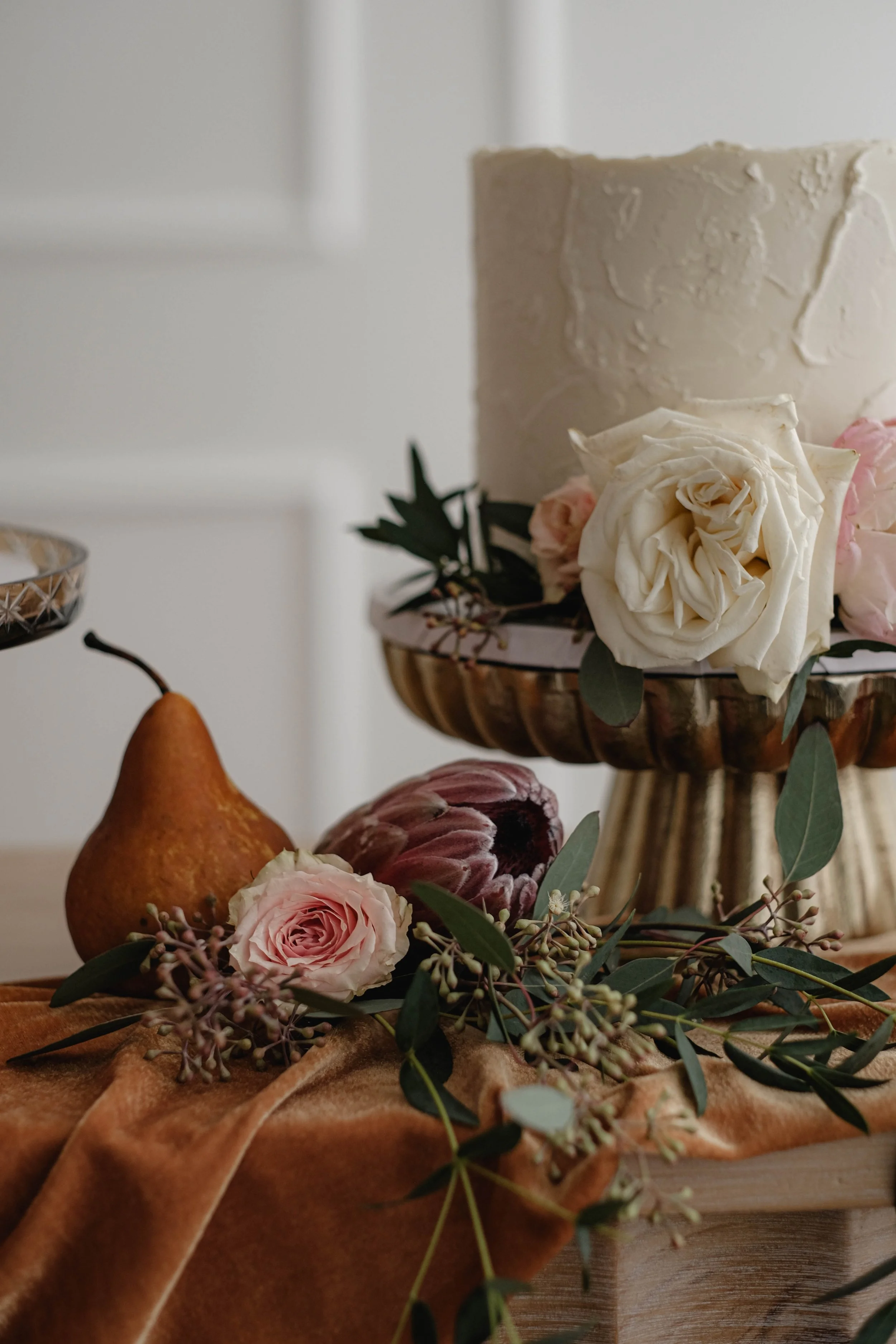 Decorative still life arrangement with a cake, flowers, and a pear on a wooden surface with an orange cloth.