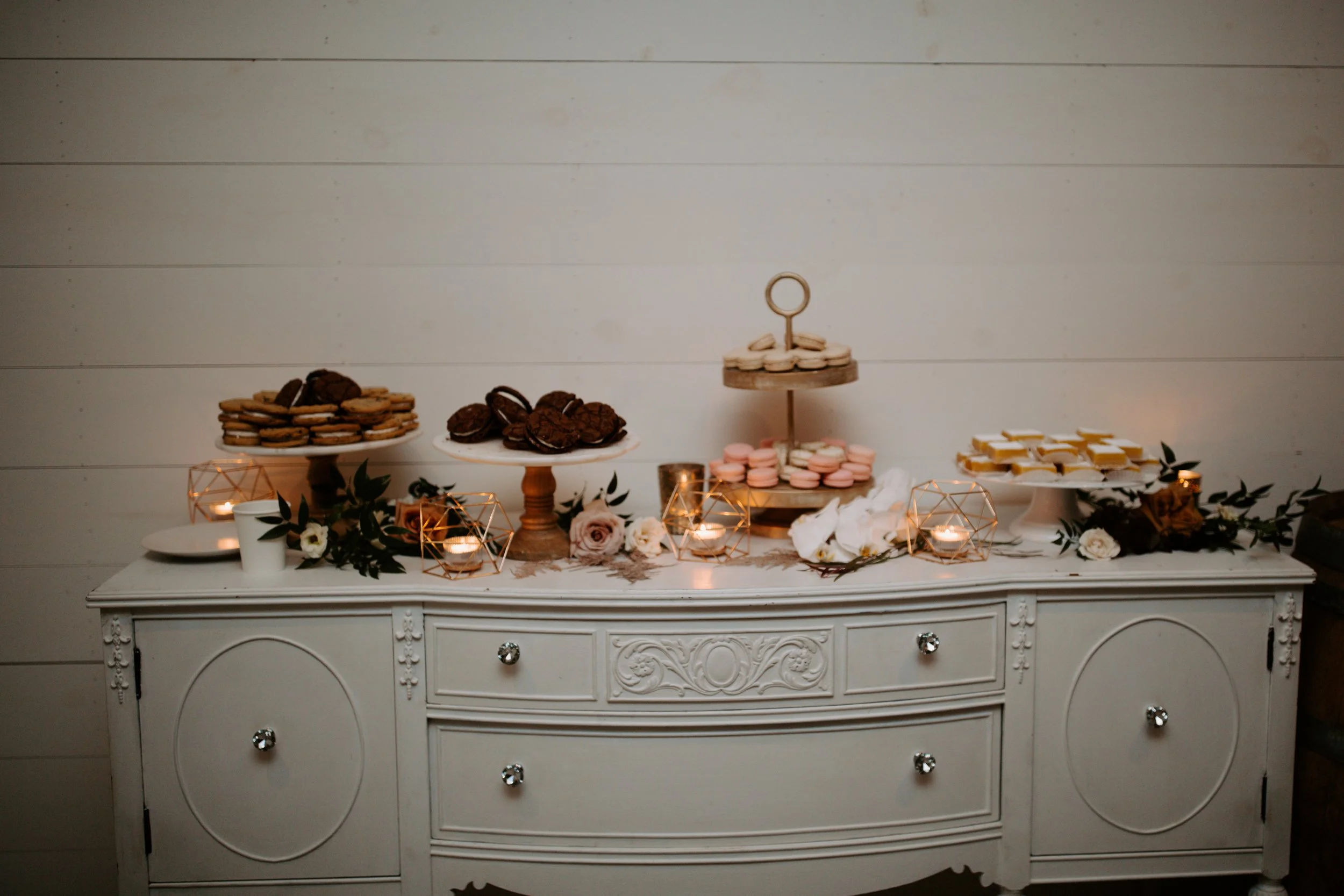Dessert table with various cookies and macarons, decorated with flowers and candle holders, against a white paneled wall.