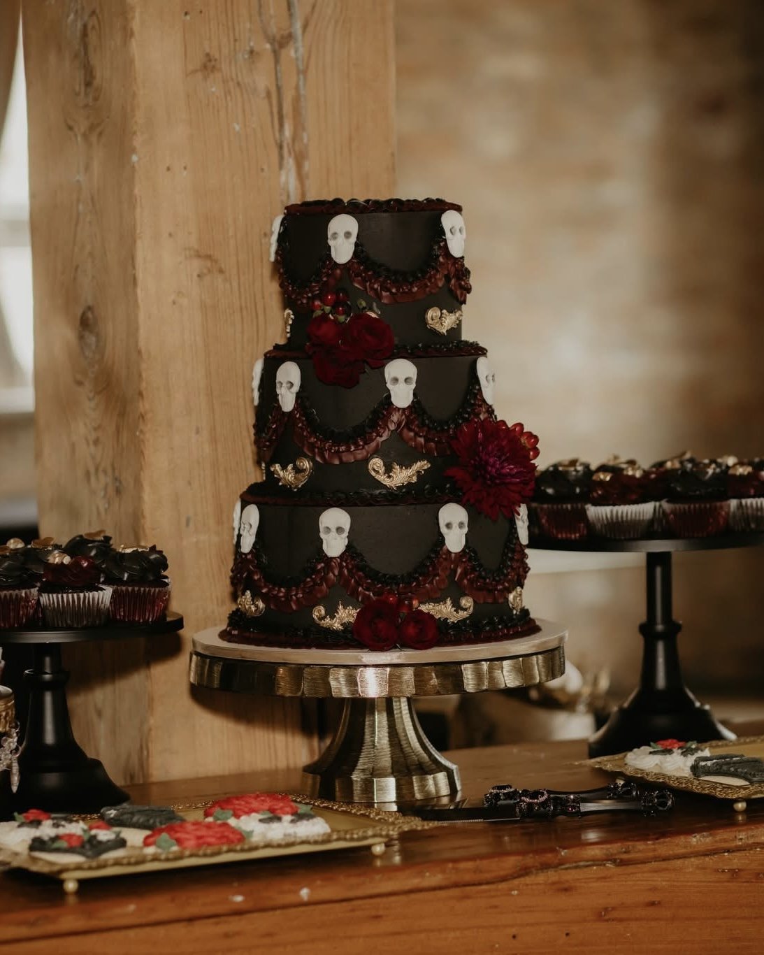 A three-tier black cake decorated with white skulls, red flowers, gold accents, and red egg-shaped candies, displayed on a metallic cake stand.