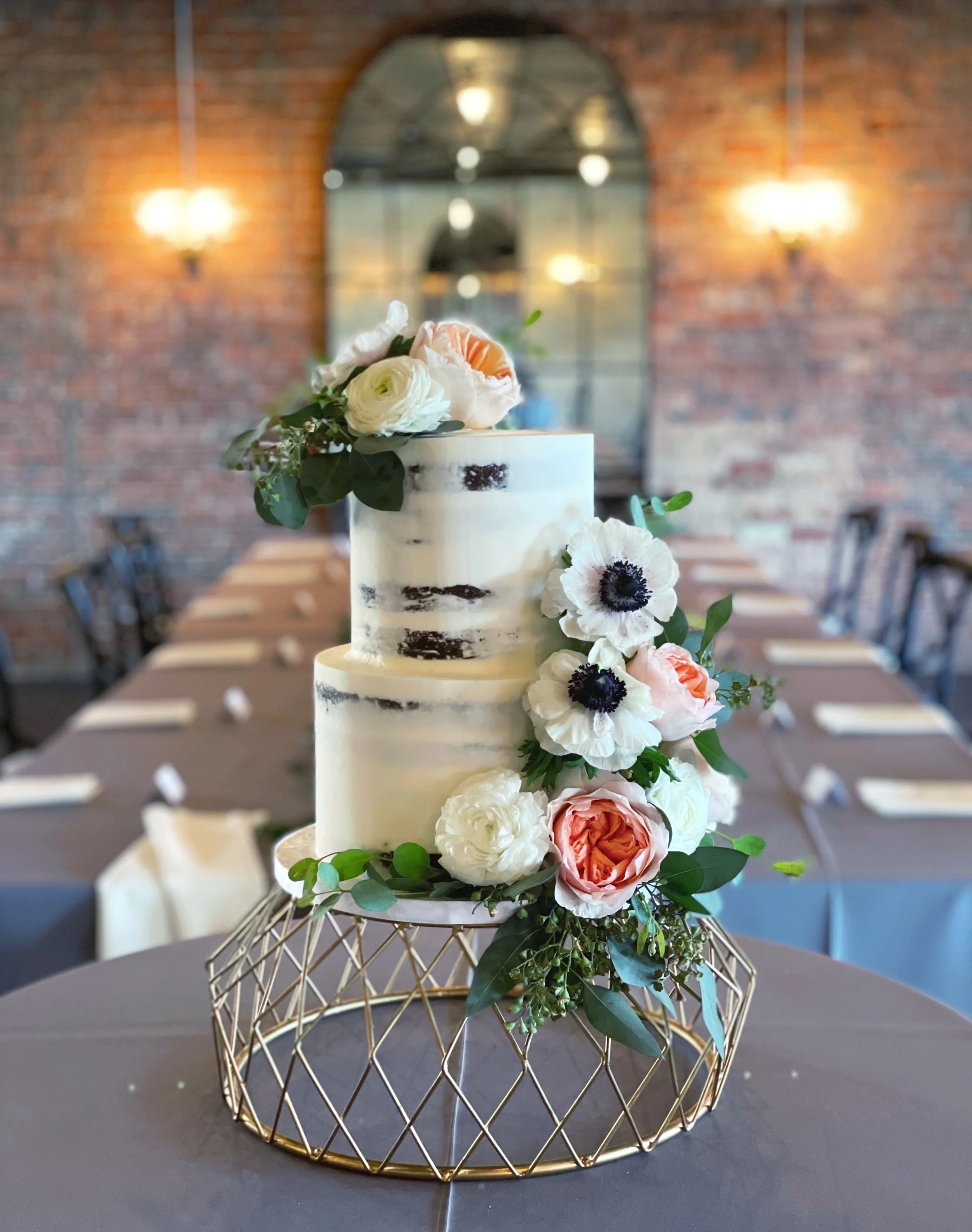 Two-tiered wedding cake with a semi-naked frosting style, decorated with white, pink, and black flowers, placed on a geometric gold stand, set on a table in a rustic wedding venue with brick walls and warm lighting.