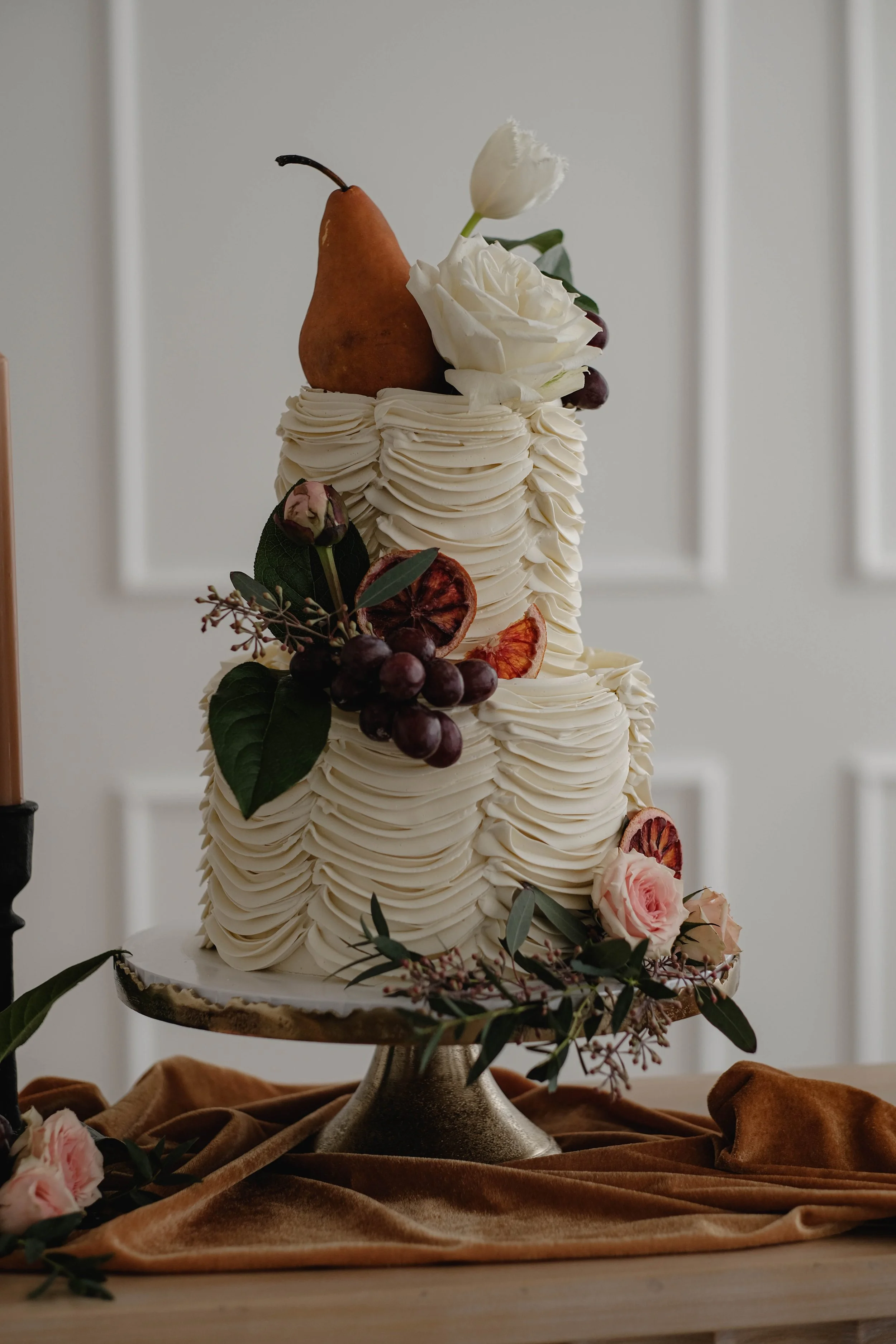 A layered wedding cake decorated with white frosting, grapes, a pear, dried fruit, roses, and greenery on a cake stand with a brown velvet cloth underneath.