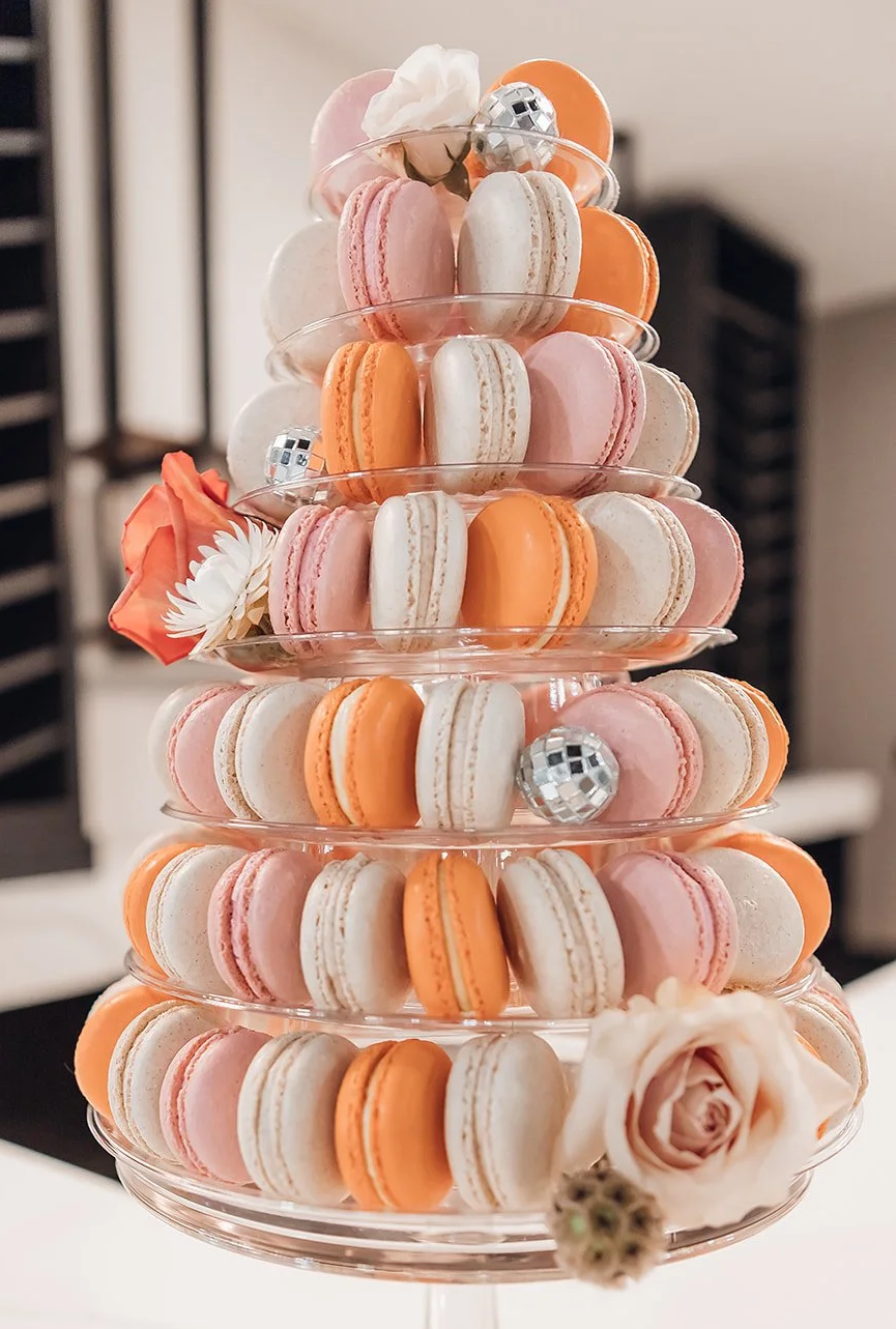 A multi-tiered glass serving stand filled with pink, white, and orange macarons, decorated with small flowers, miniature disco balls, and a large peach-colored rose.