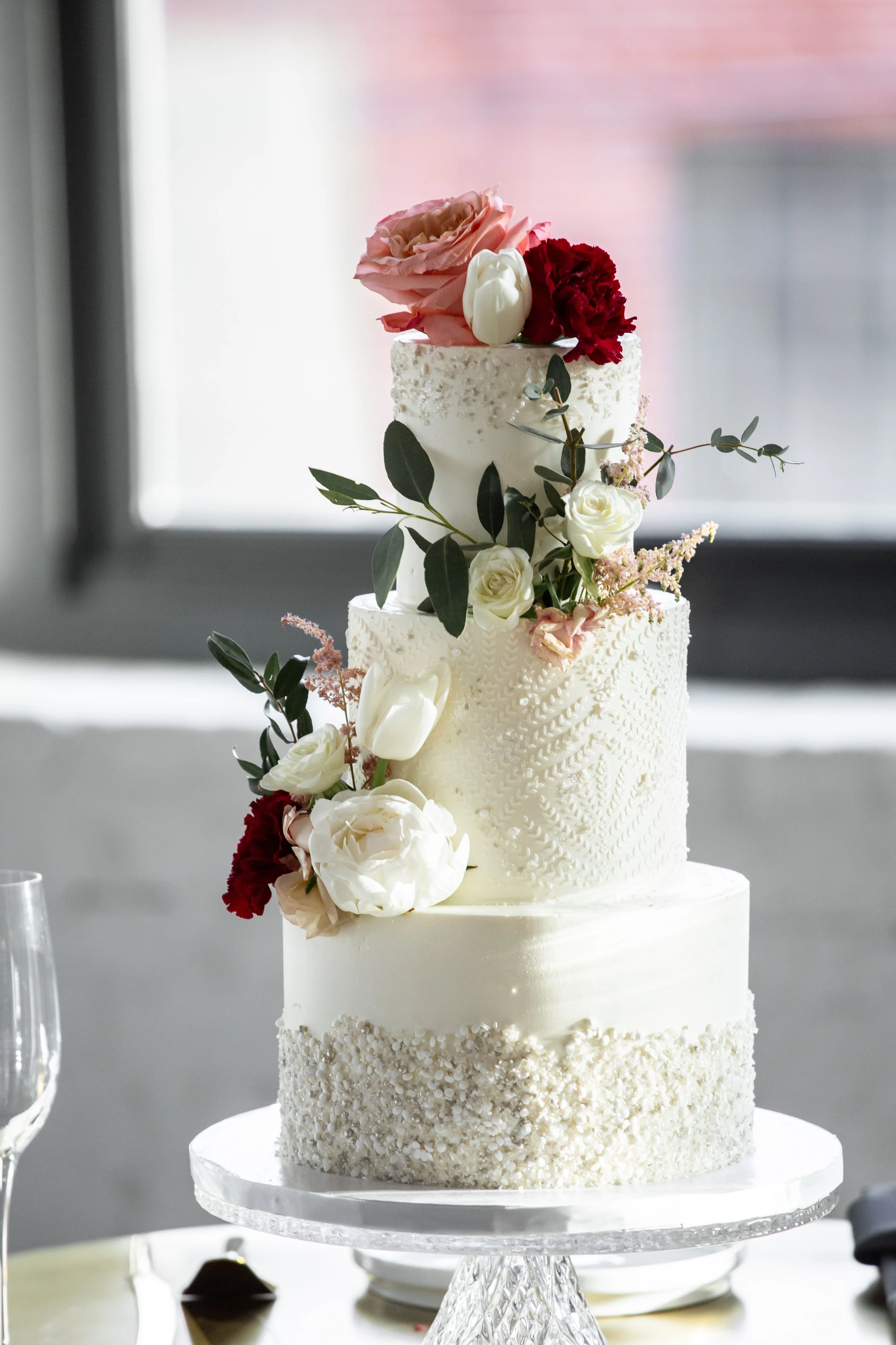 Elegant four-tier white wedding cake decorated with white and red flowers, green leaves, and small pink accents, placed on a glass cake stand.