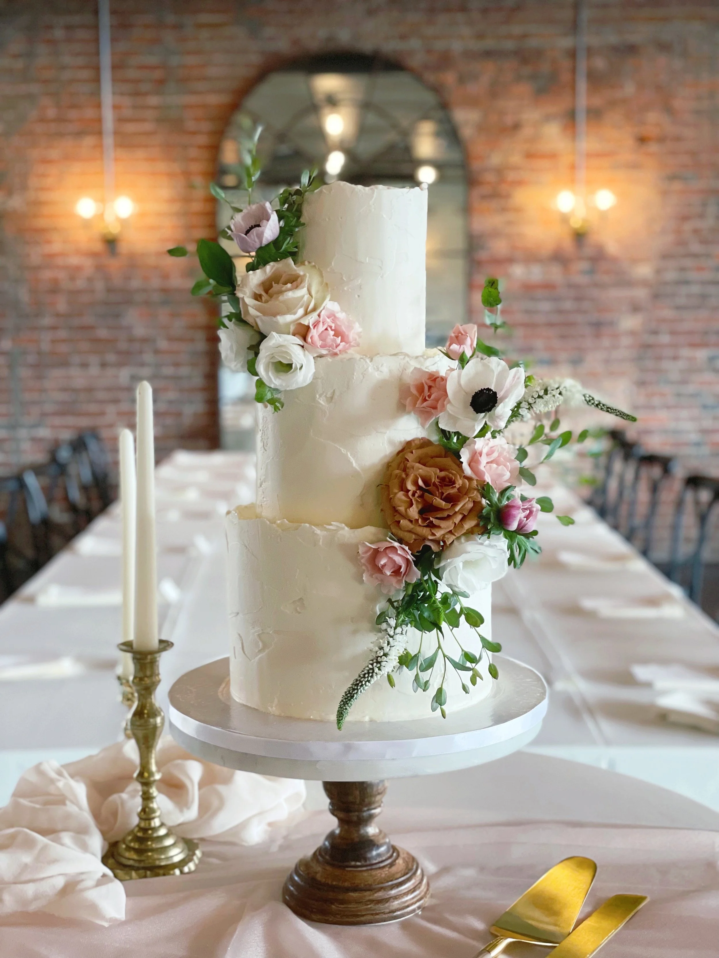A three-tier wedding cake decorated with pastel pink, white, and brown flowers, placed on a white cake stand in a decorated venue with a brick wall and mirror in the background.