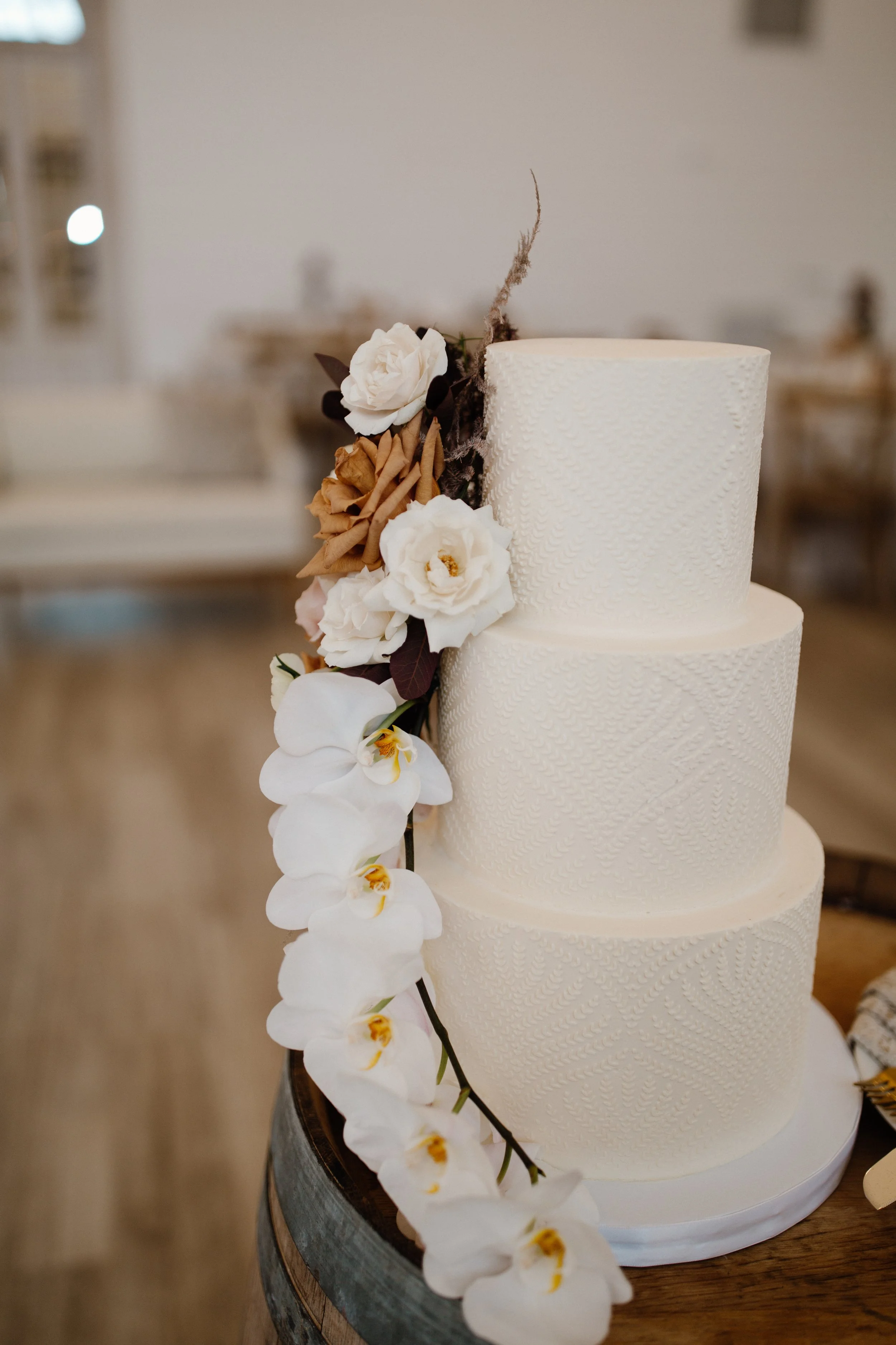 A tall, white, three-tier wedding cake with textured icing, decorated with white roses, orchids, and brown flowers on the side, placed on a wooden stand.