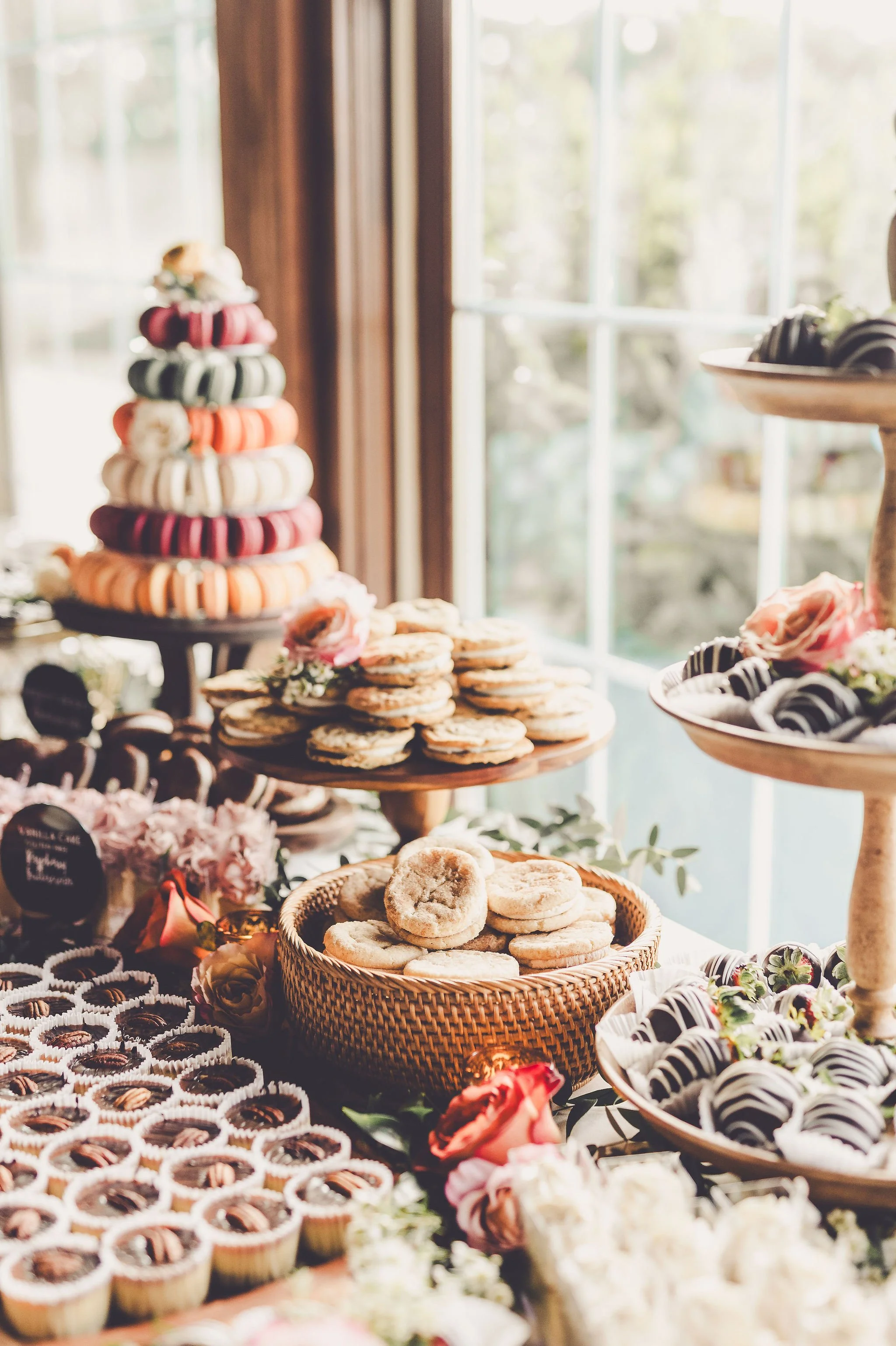 A dessert table with assorted cookies, macarons, and chocolates arranged on tiered trays and in baskets near a window with sunlight.