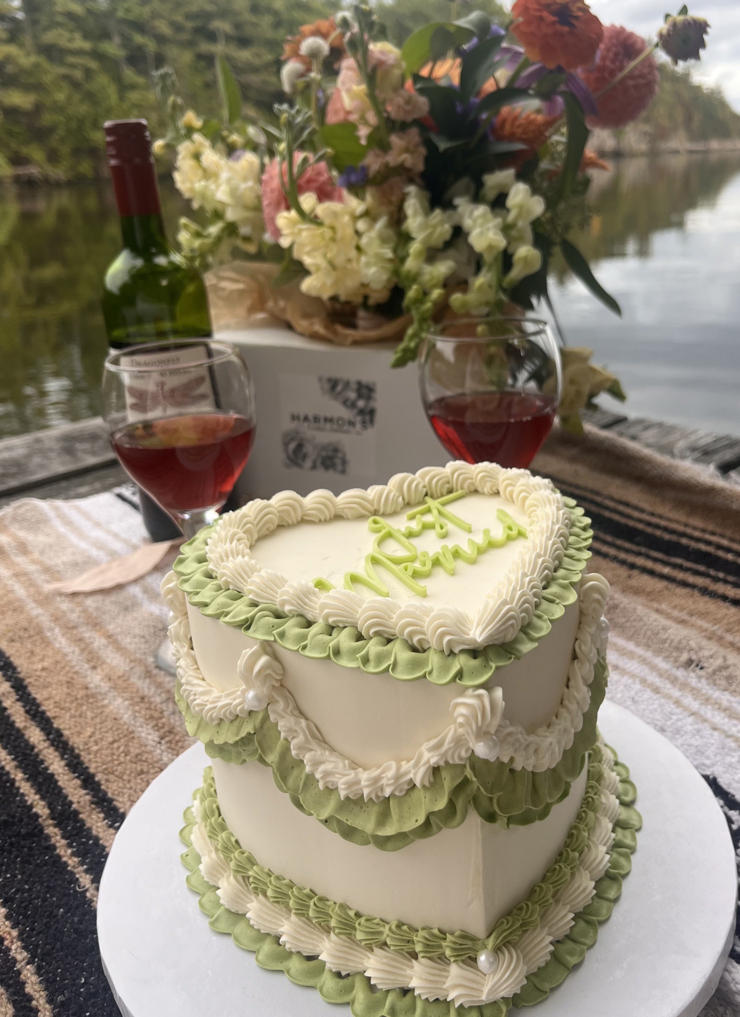 A heart-shaped wedding cake with white, green, and cream frosting, decorated with piped icing and a green inscription on top. Two glasses of rosé wine, a bottle of wine, and a large bouquet of flowers in a white box are on a table by a river, with tr
