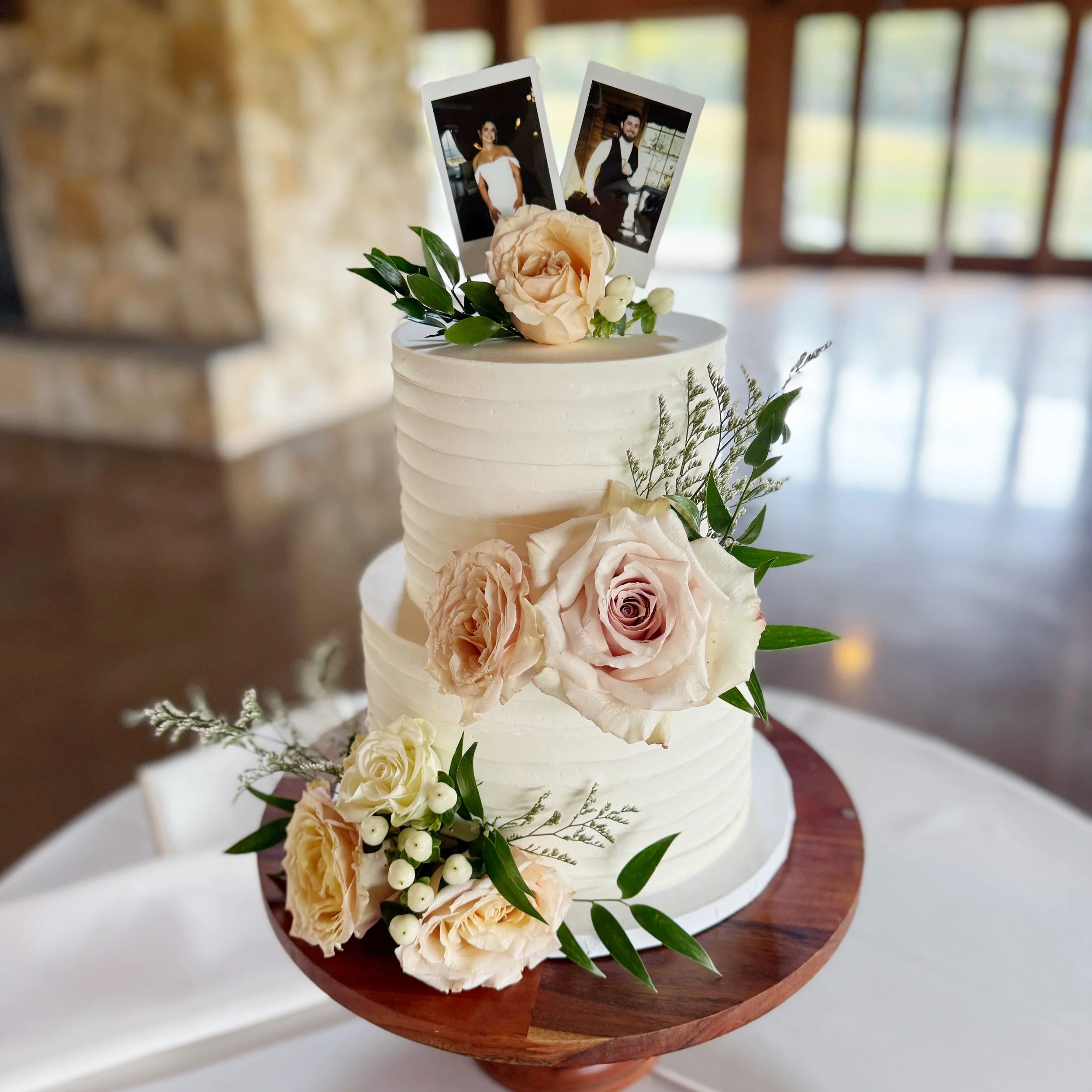 A two-tier white wedding cake decorated with pink and white roses, greenery, and small white berries, topped with two Polaroid photos of a bride and groom, on a wooden cake stand.
