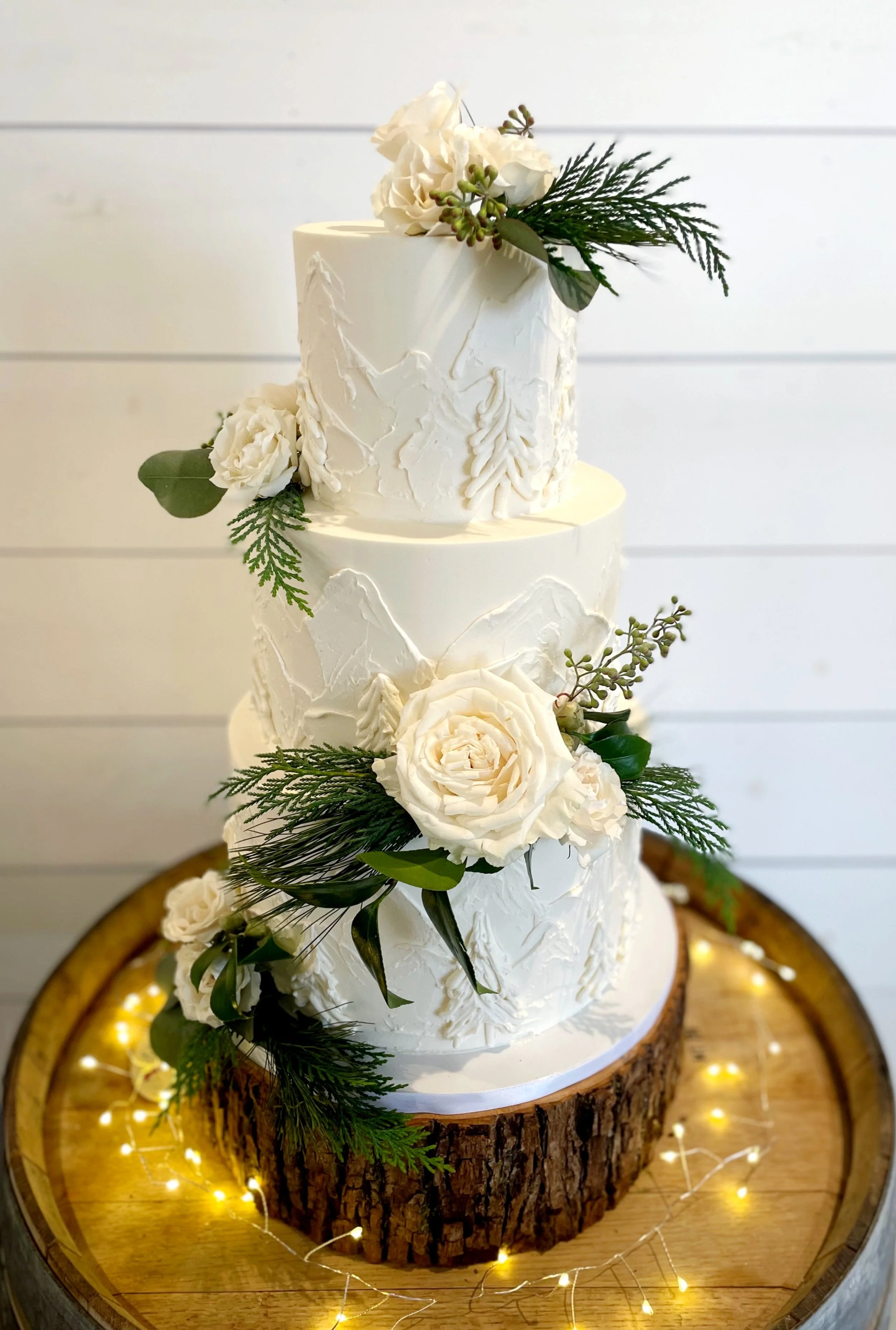Three-tier white wedding cake decorated with white flowers and greenery, placed on a wooden slab with fairy lights around the base.