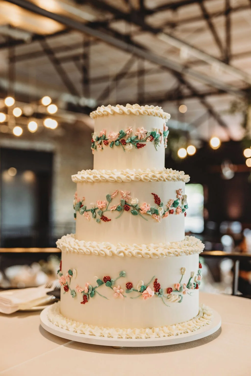 A four-tiered wedding cake with white icing, decorated with pastel-colored floral piping and green leaves, set on a table in a venue with exposed beams and warm lighting.