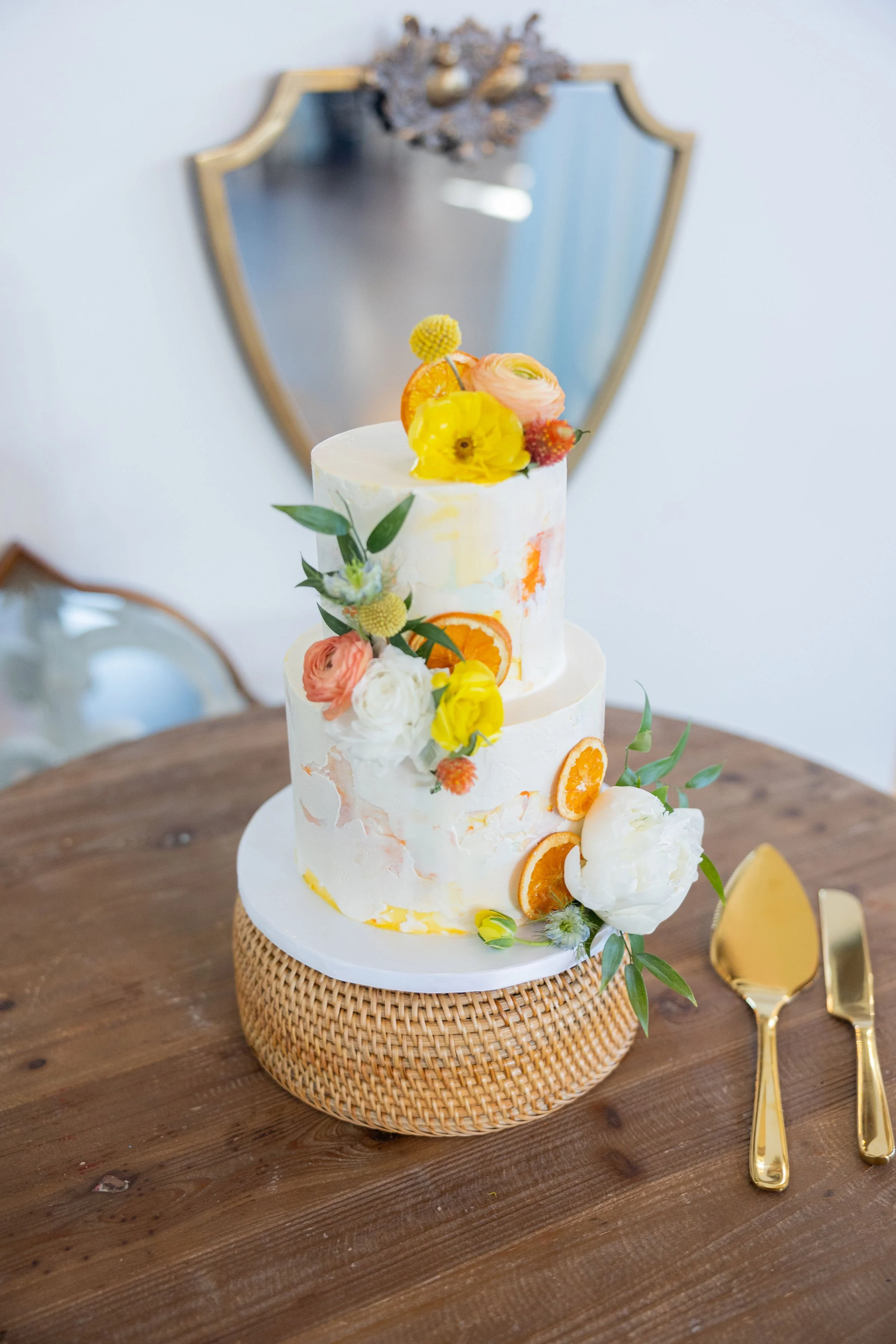 Two-tier white wedding cake decorated with orange slices and fresh flowers, placed on a wooden table with gold utensils nearby.