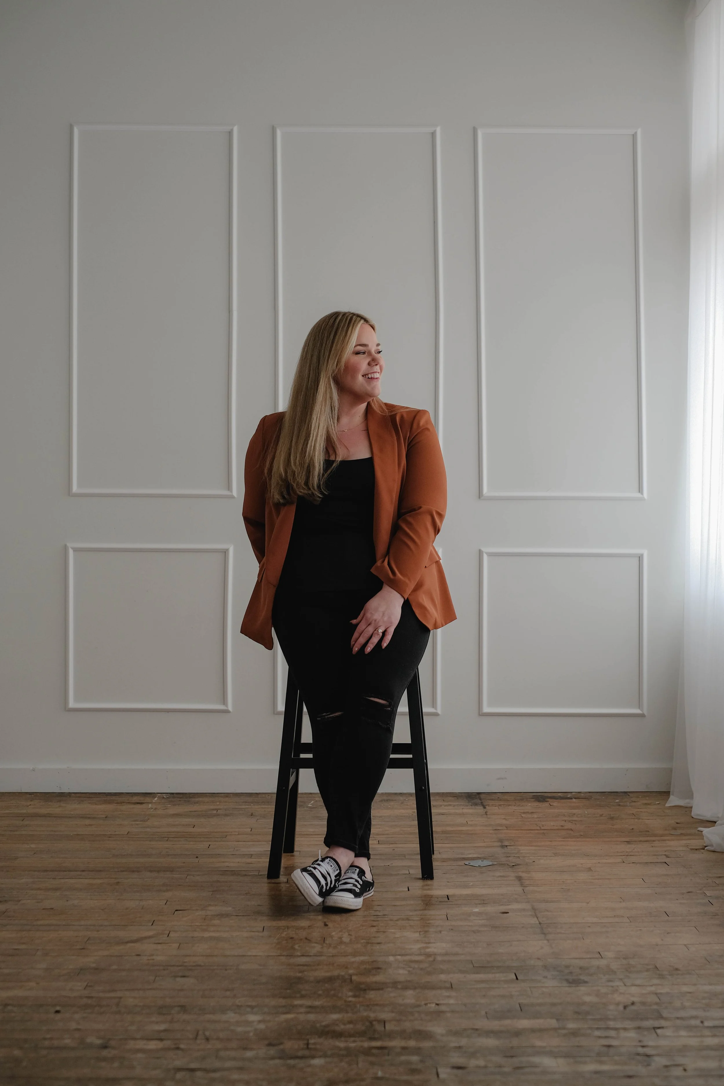 A woman with long blonde hair leaning against a black chair in a minimally decorated room with white paneled walls and hardwood floor, smiling and looking to her right.