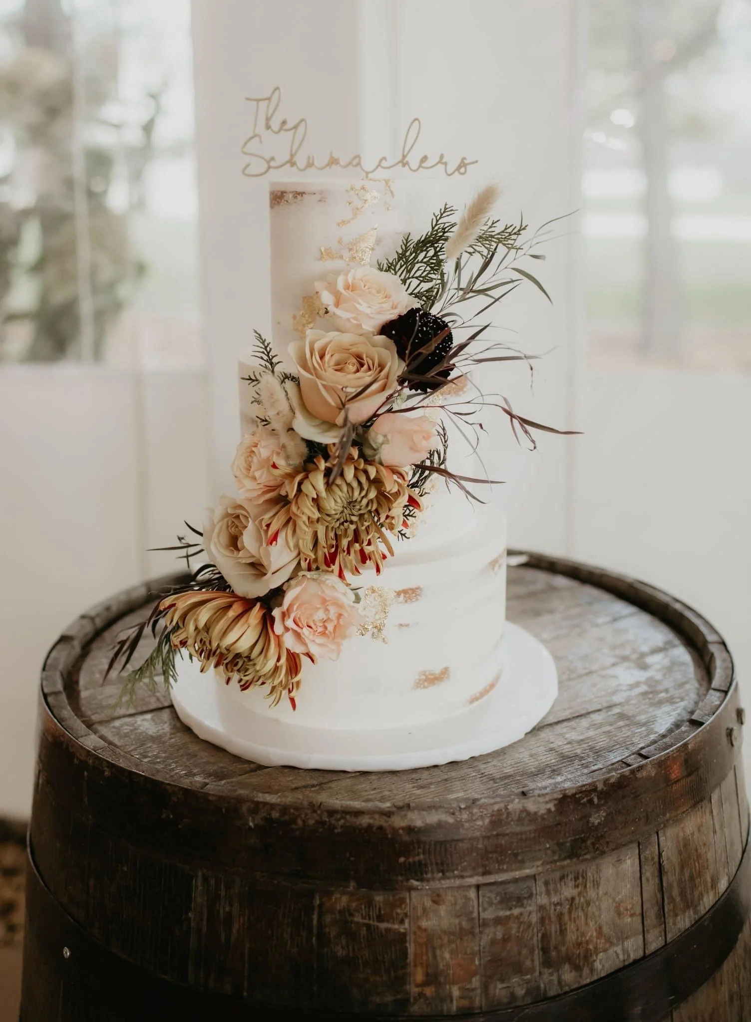A white, semi-naked wedding cake decorated with peach and blush flowers, greenery, and gold accents, sitting on a wooden barrel.