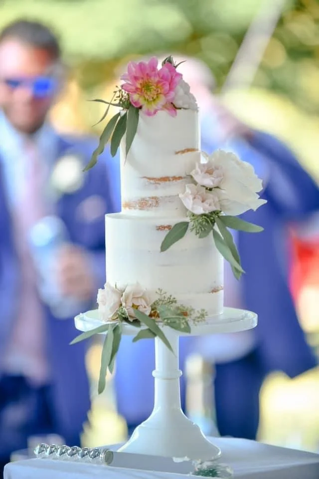 A two-tier white wedding cake decorated with pink and white flowers and green leaves on a white cake stand at an outdoor event.