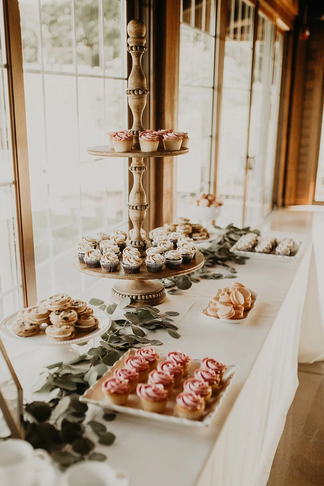 A dessert table with cupcakes, cookies, and macarons, decorated with eucalyptus leaves, in a room with large windows and wooden framing.