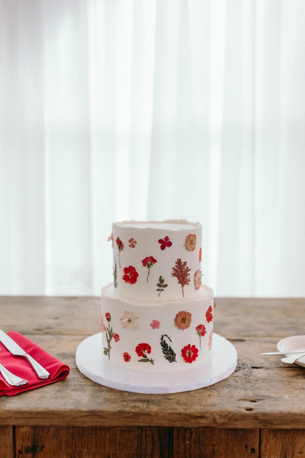 A two-tiered white cake decorated with edible flowers and botanical patterns, placed on a wooden table with a red napkin and silverware on the side.
