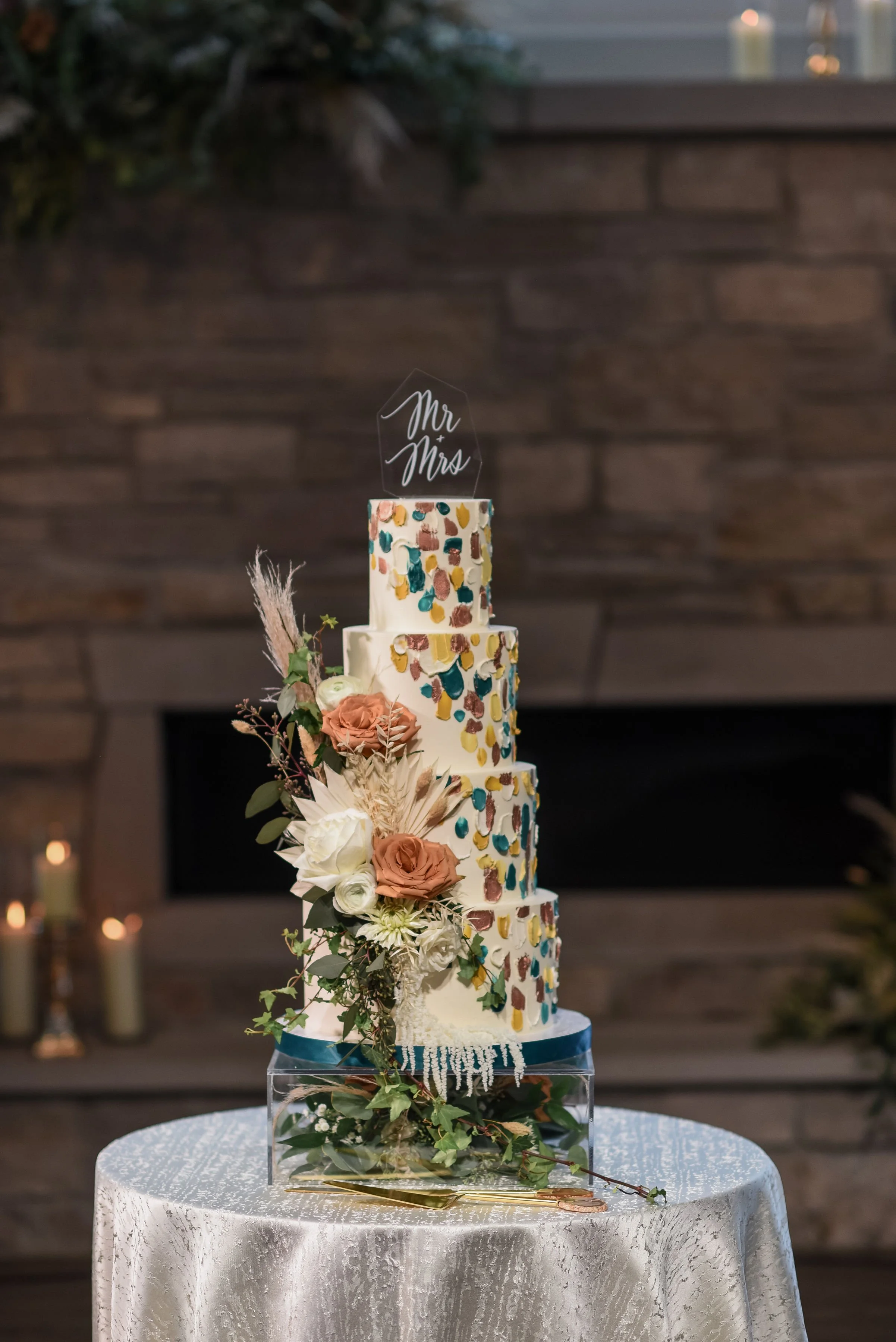 Four-tiered wedding cake decorated with colorful brushstroke patterns and fresh flowers, topped with a acrylic cake topper that says 'Mr & Mrs', on a table with a silver tablecloth.