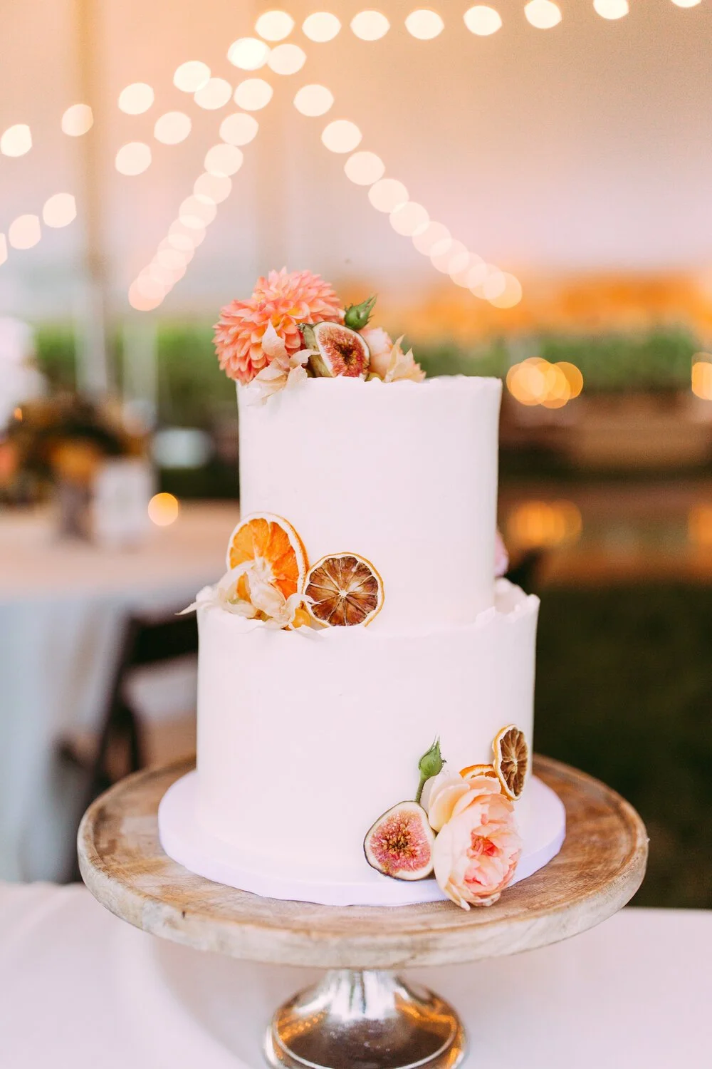 Two-tier white wedding cake decorated with dried citrus slices, peaches, and pink flowers, placed on a wooden cake stand.