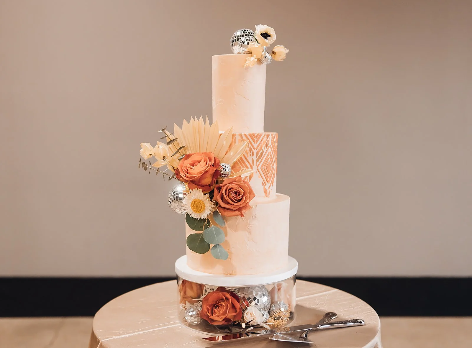 Three-tiered wedding cake with pastel pink and peach frosting, decorated with fresh roses, daisy, eucalyptus leaves, and silver disco balls, on a round table with cake-cutting utensils.