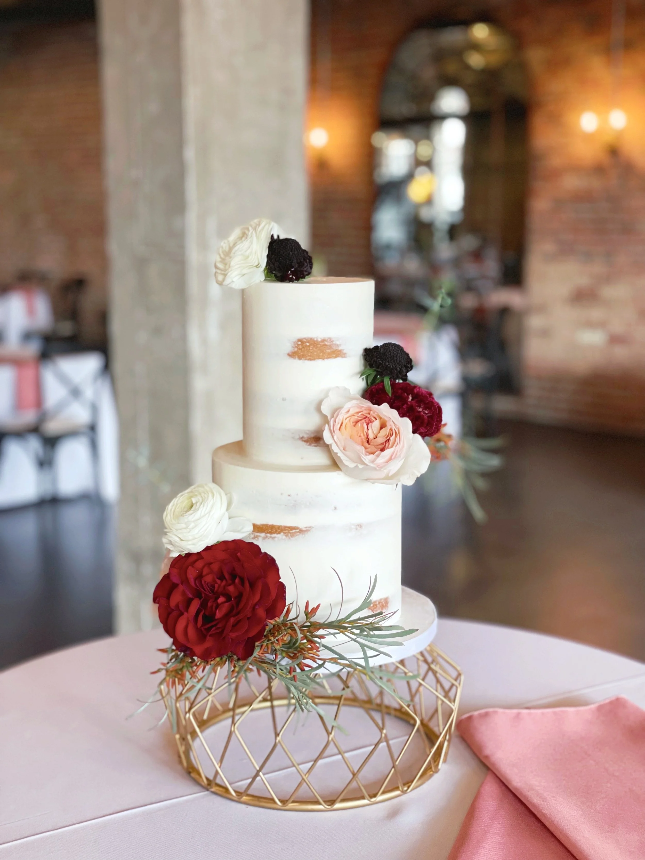 Two-tier white wedding cake decorated with dark red, blush, and black flowers, placed on a decorative gold stand with a pink cloth napkin nearby in a rustic indoor setting.