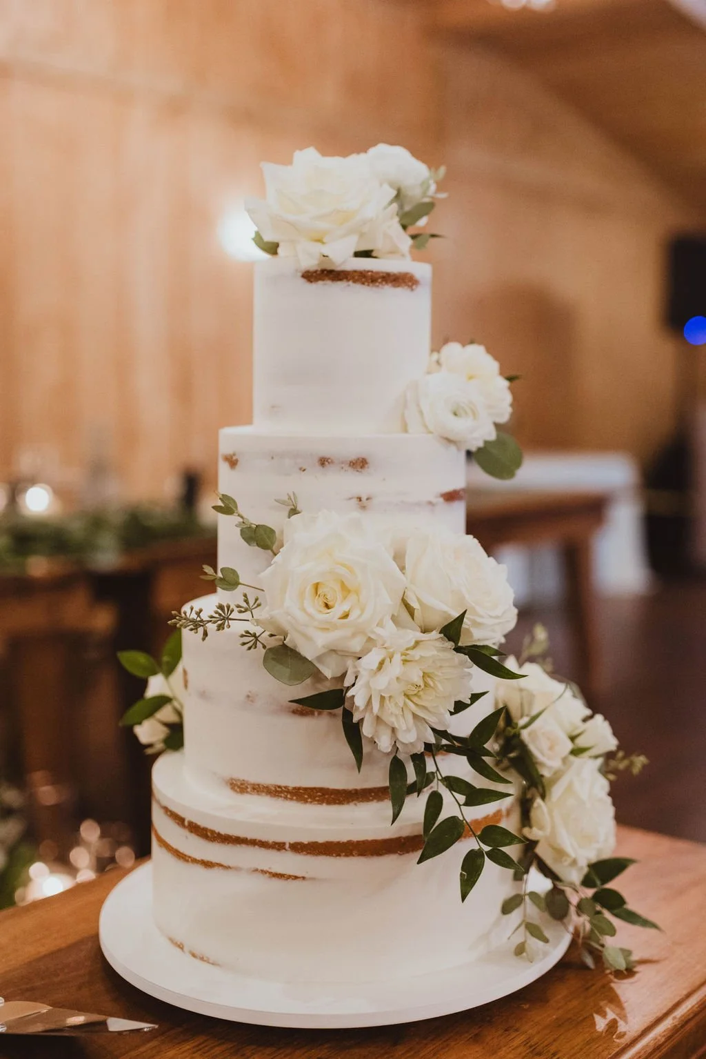 Three-tier wedding cake with white frosting, decorated with white roses and green foliage, on a wooden table in a warmly lit room.