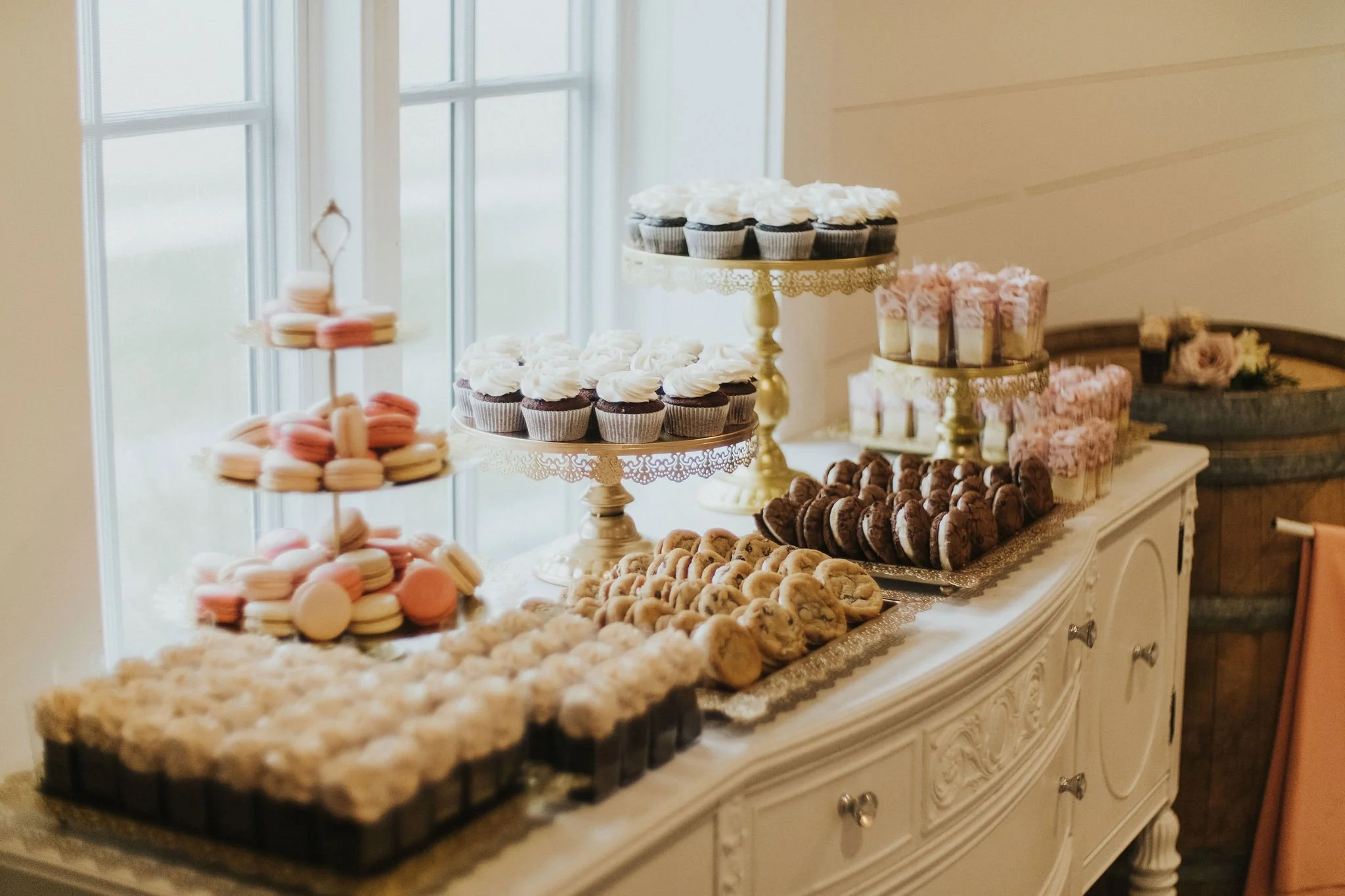 A dessert table with assorted cupcakes, cookies, macarons, and pastries on elegant stands and trays near a window.