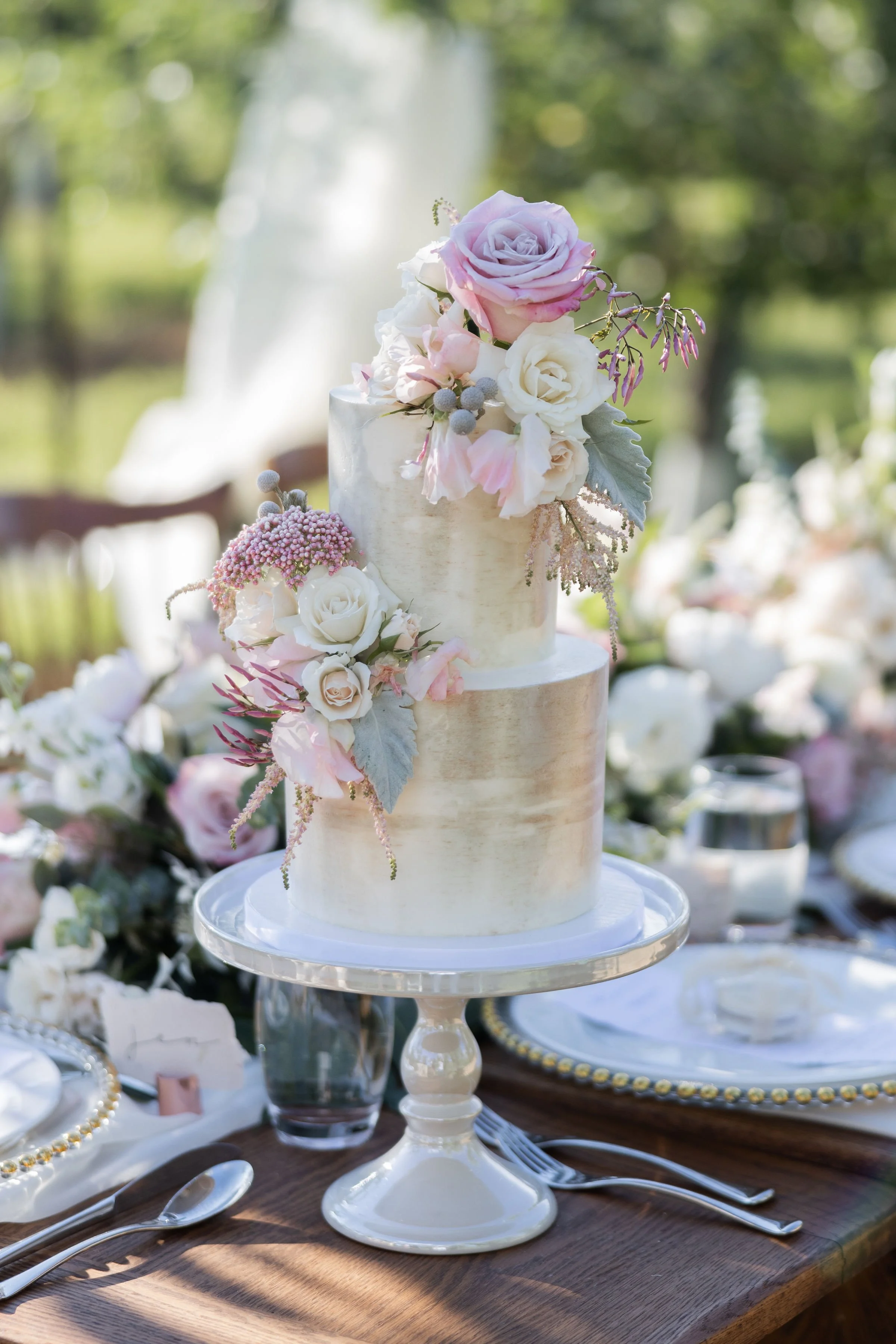 Elegant wedding cake with two tiers, decorated with pink and white roses, greenery, and purple accents, placed on a white cake stand on a wooden table outdoors.