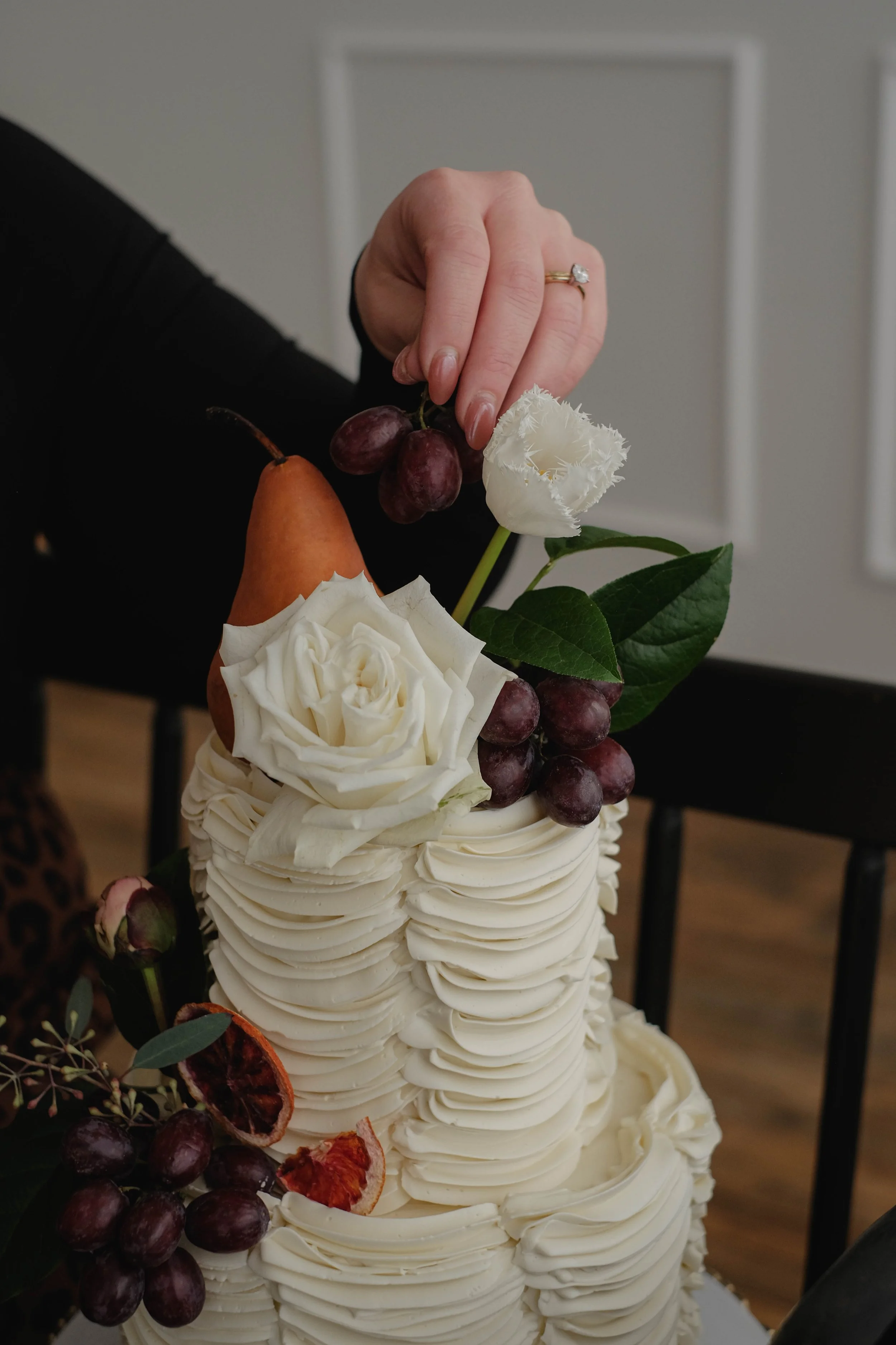 Close-up of a wedding cake decorated with grapes, orange pear, white roses, and white flowers, with a person's hand adjusting the floral decoration.