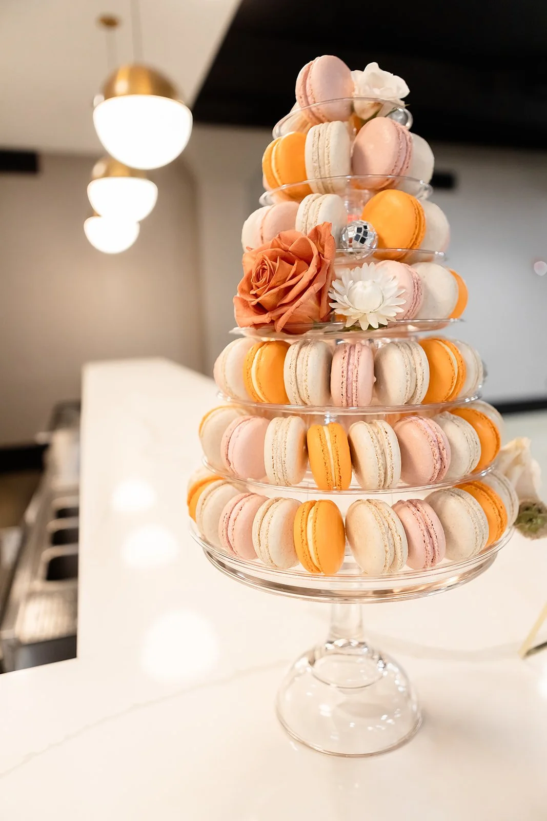 A tiered glass stand filled with pink, white, and orange macarons, decorated with a pink rose, a white flower, and a silver disco ball ornament.
