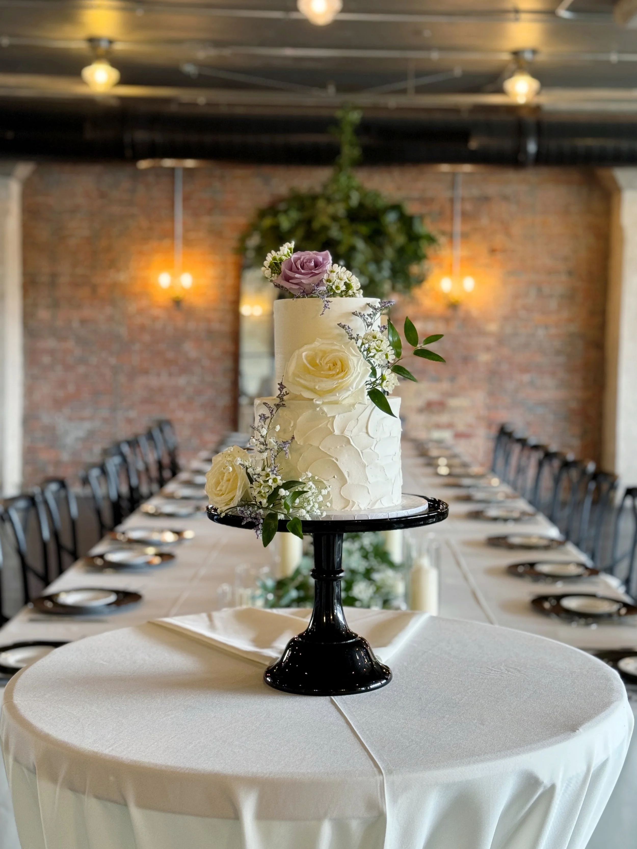 A wedding cake with white icing decorated with white and purple flowers on a black cake stand, placed on a round table with a white tablecloth.