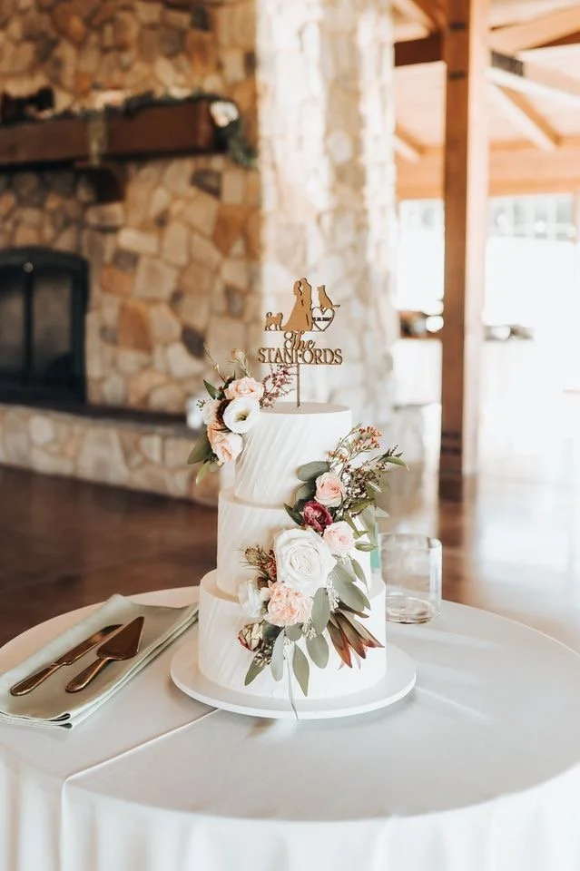 A wedding cake decorated with pink and white flowers and green foliage, with a topper that says 'The Stanfords' and has a silhouette of a dog, set on a white tablecloth-covered table.