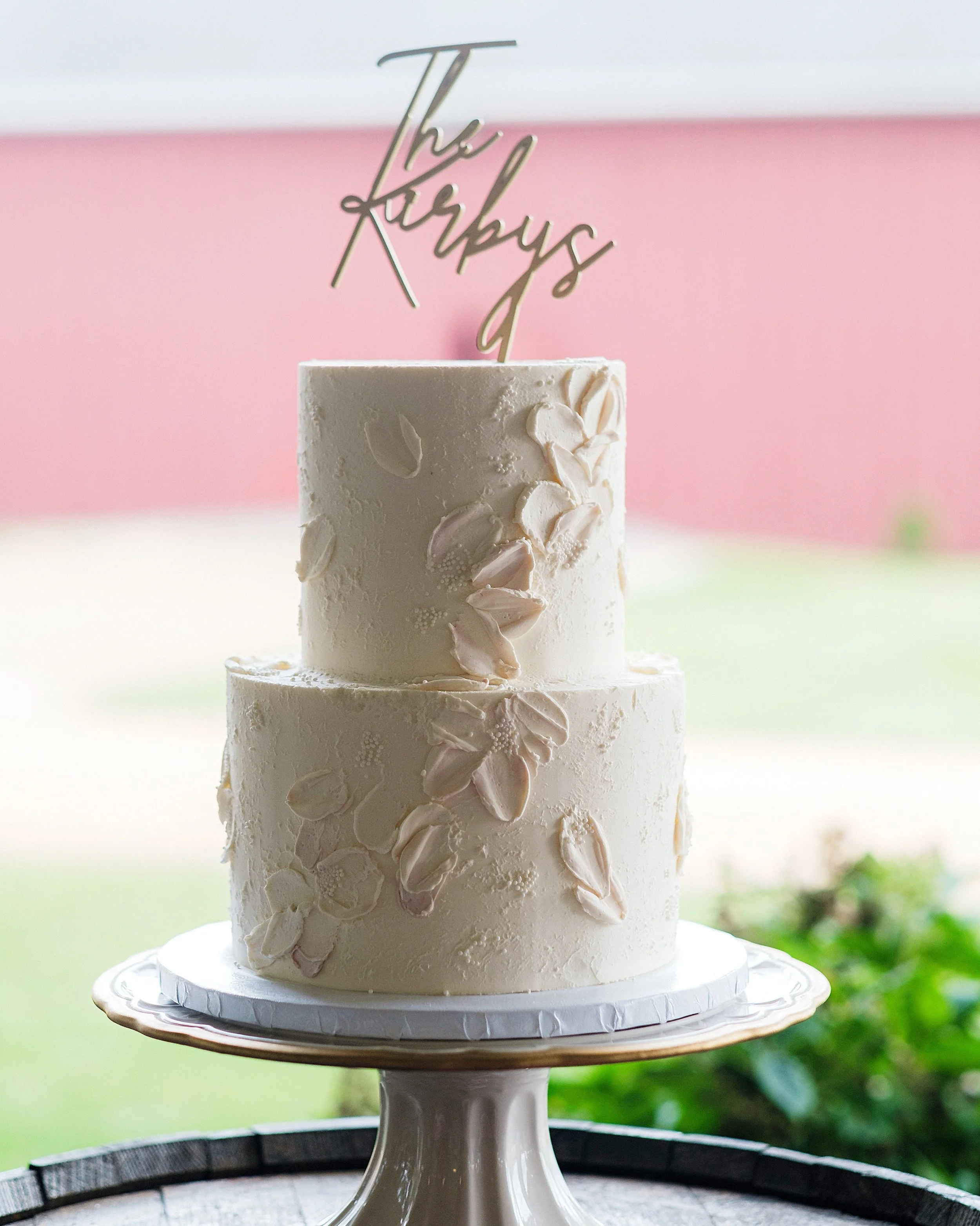 Two-tiered white wedding cake with almond slices, topped with a gold stick cake topper that says 'The Karbys'.