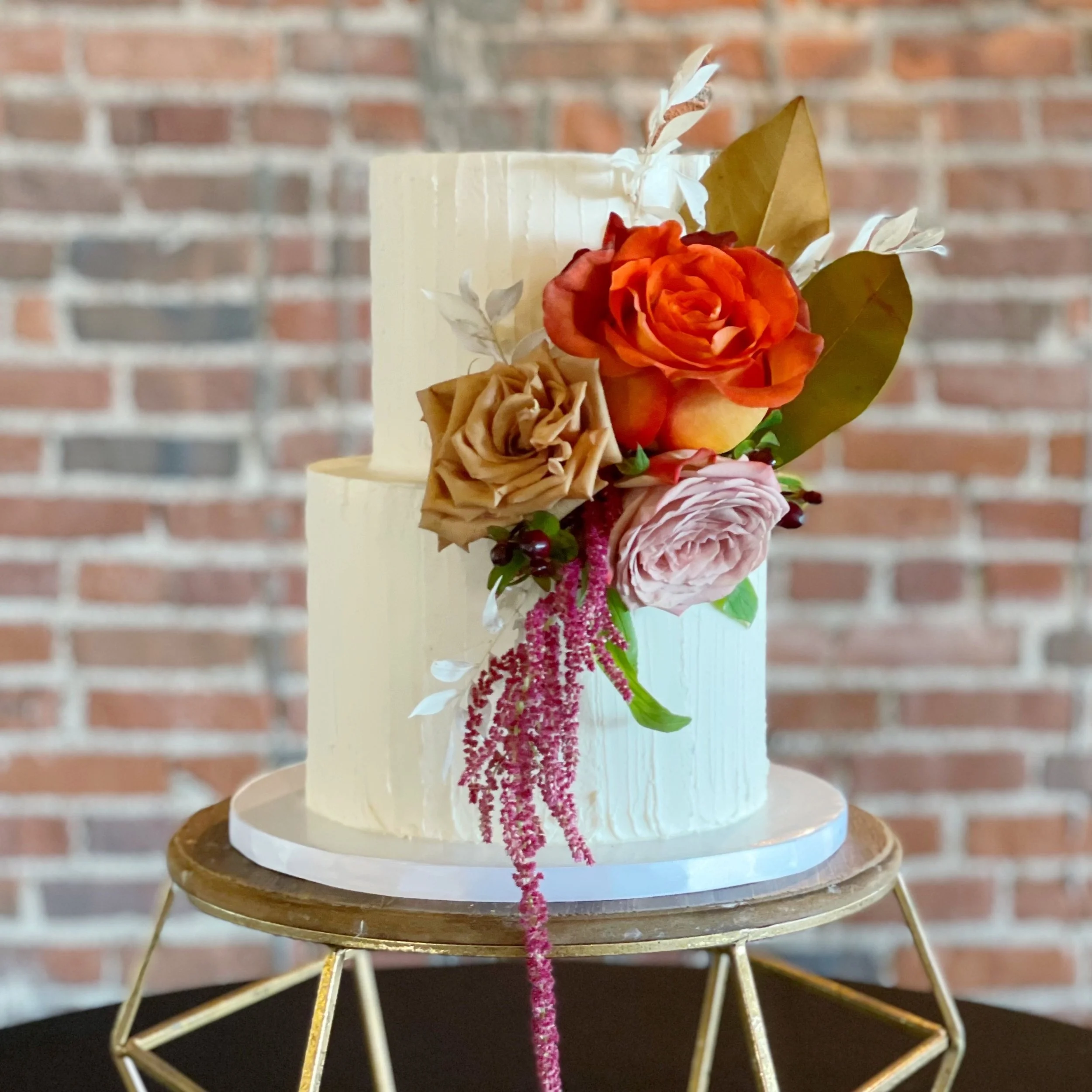 A two-tier cream-colored wedding cake with floral decoration on top, featuring orange, beige, and pink roses, green leaves, and trailing pink flowers, placed on a wooden cake stand against a brick wall background.