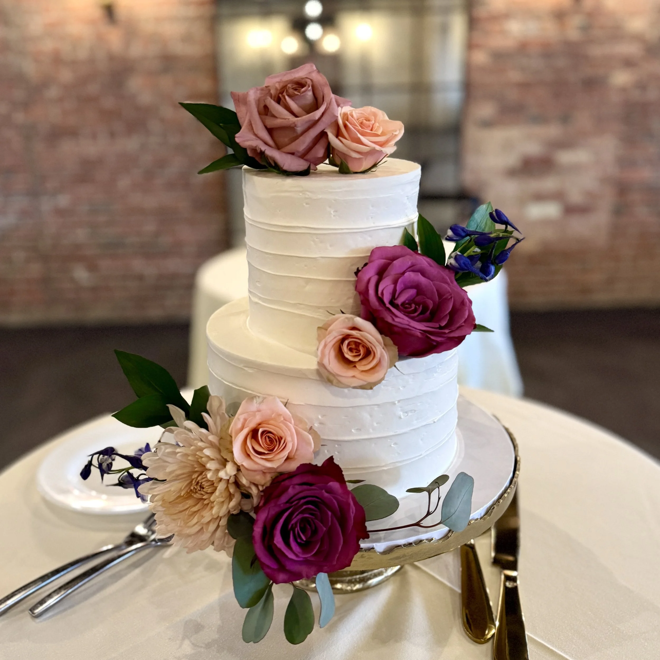 Two-tiered white wedding cake decorated with pink, purple, and peach roses and greenery, placed on a gold cake stand on a white tablecloth, with cake knives and a fork beside it.