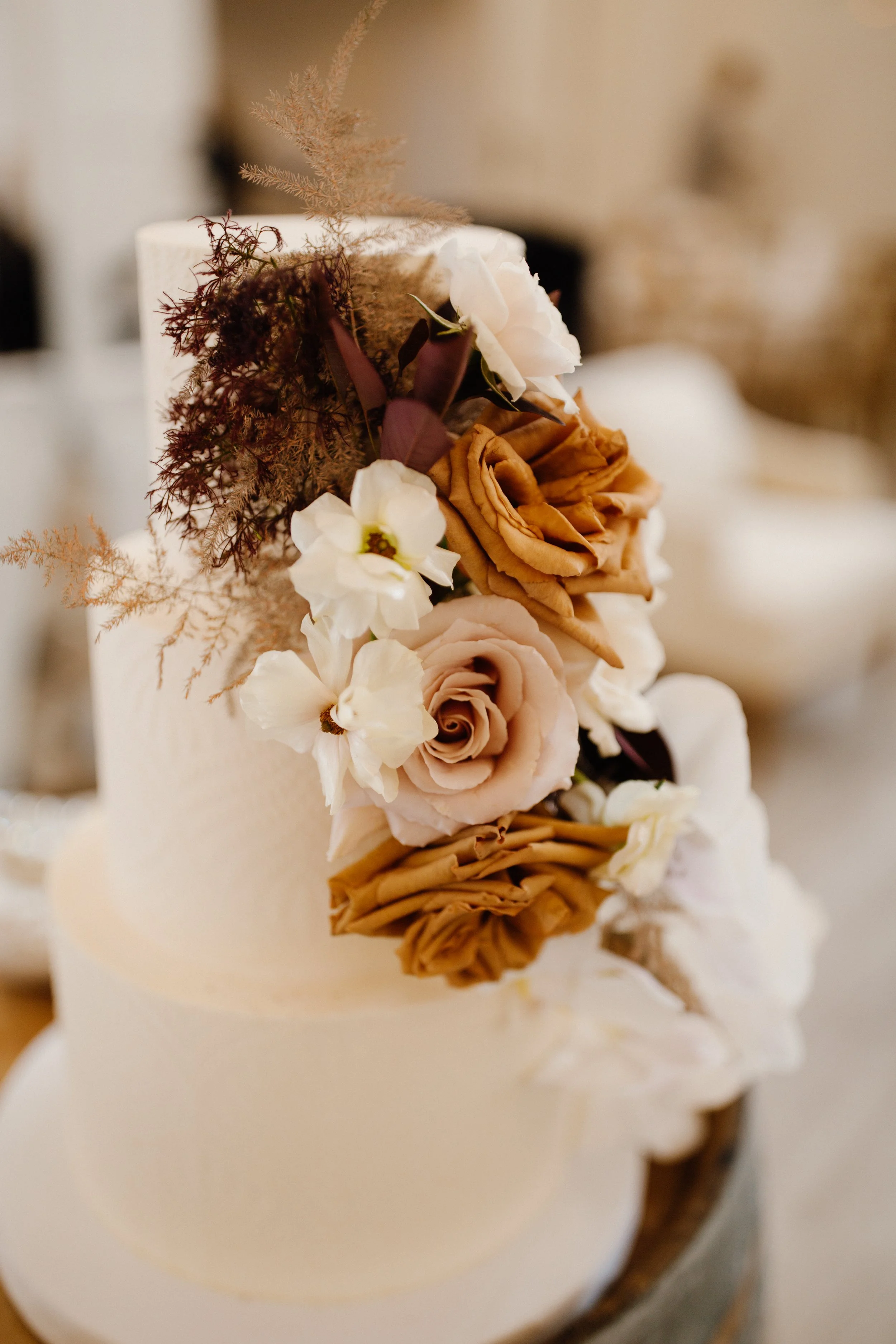 A white wedding cake decorated with a cascading arrangement of beige, brown, and white roses, complemented by dried foliage and leaves.