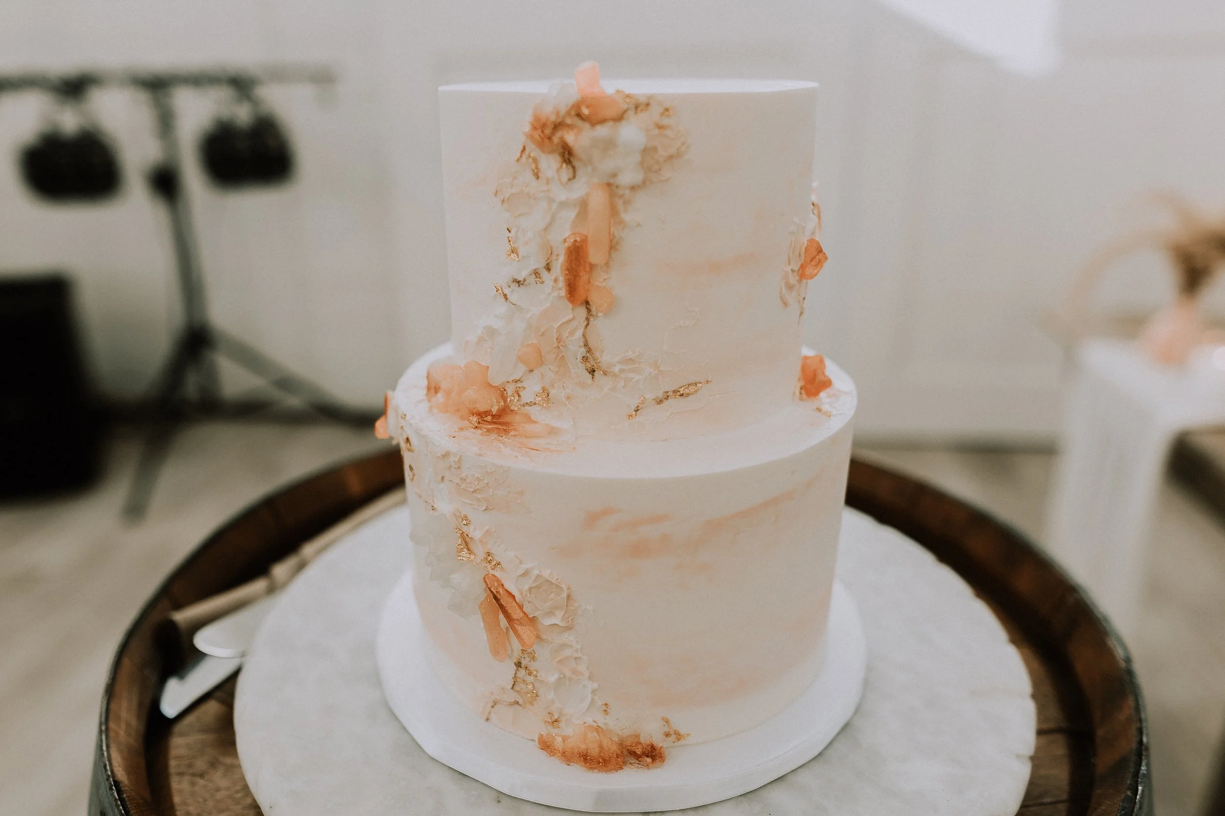 A two-tiered white wedding cake with peach-colored accents and gold leaf details, decorated with abstract icing adornments, placed on a wooden tray.
