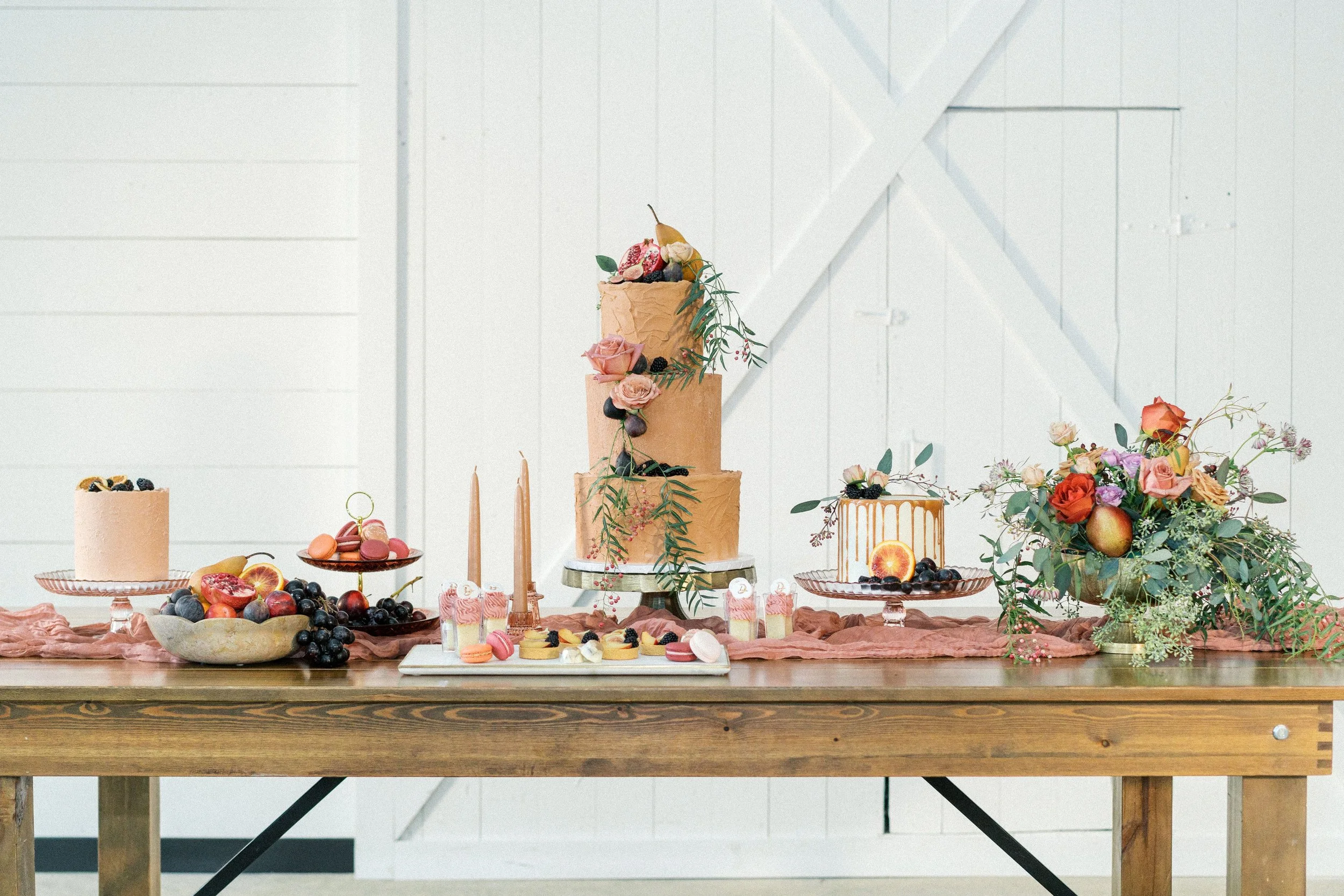 Decorative dessert table with a three-tiered cake decorated with pink roses, greenery, and fruits, along with various smaller cakes, macarons, and fresh fruit, all arranged on a rustic wooden table with floral arrangements in a white vase and pink fa