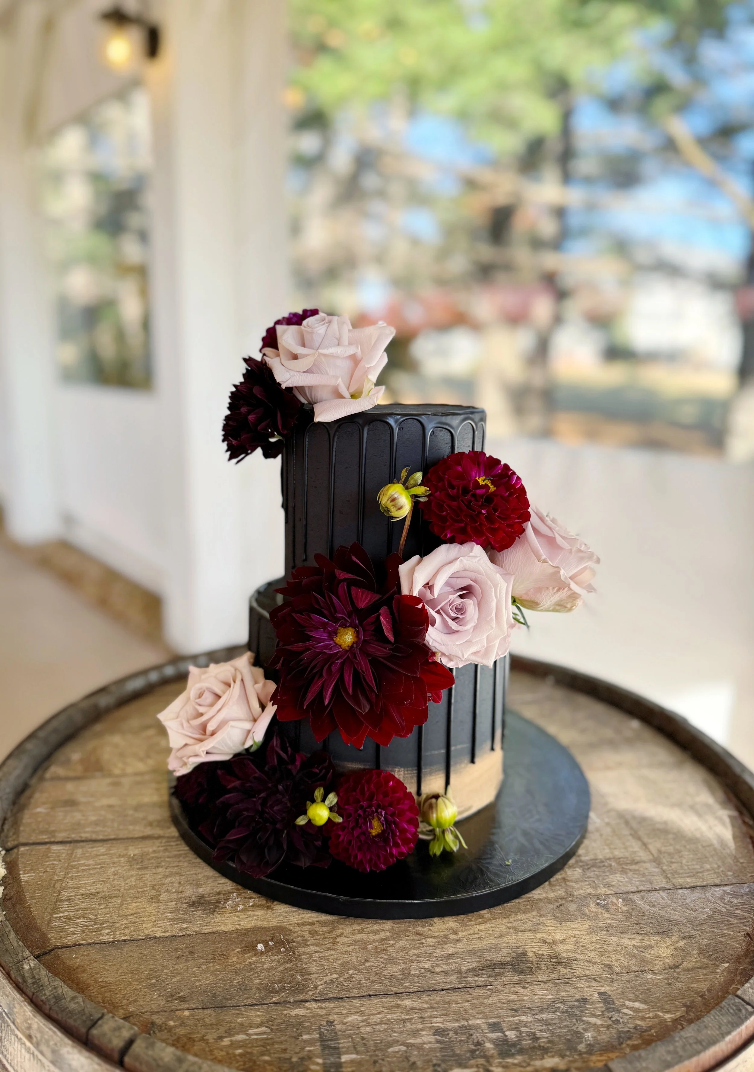 A tall, black, rectangular cake decorated with light pink roses, deep burgundy dahlias, and small buds, placed on a round wooden table with rustic planks, with a background of a porch and trees outside.