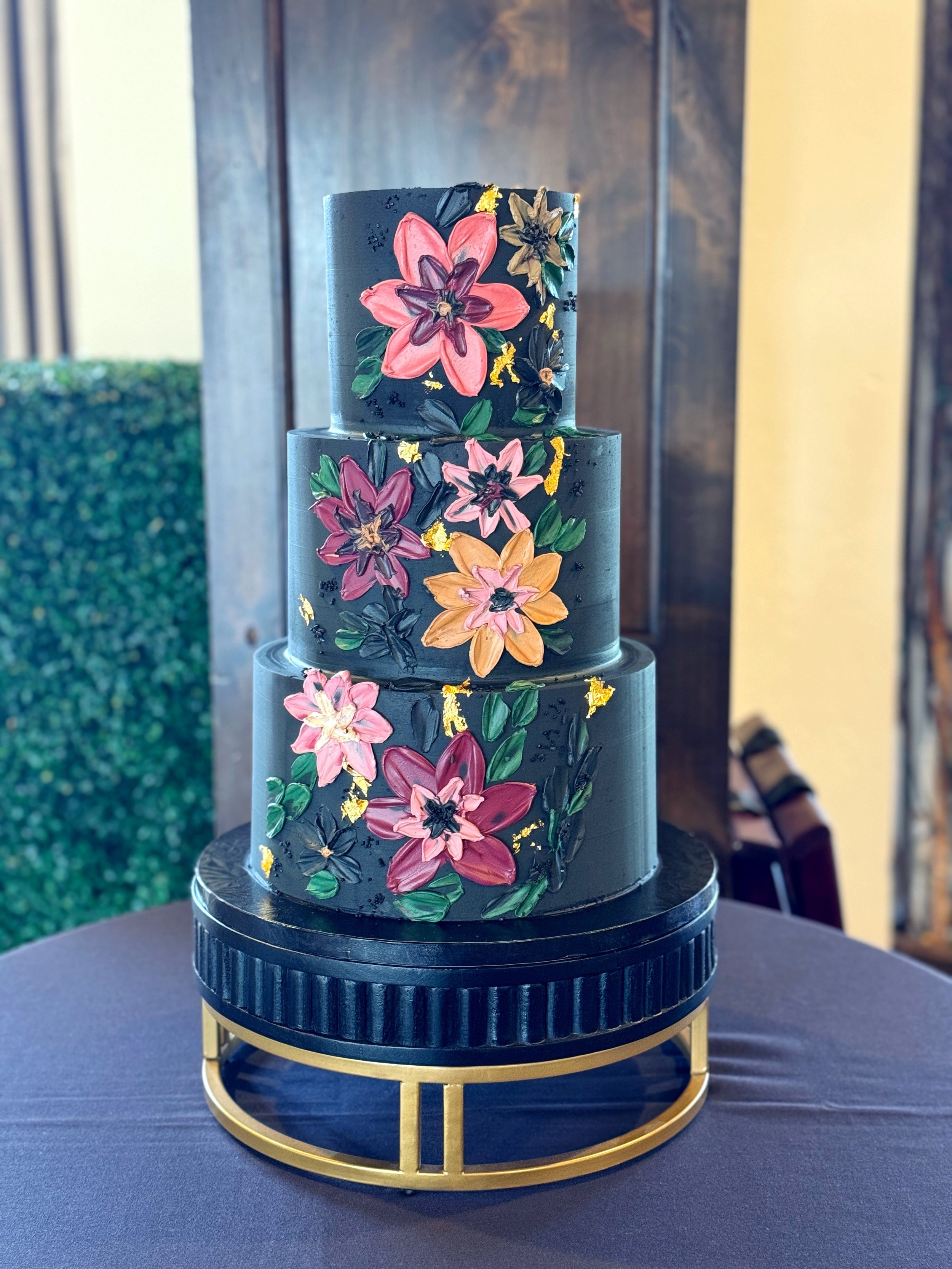 A three-tiered black cake decorated with colorful hand-painted flowers and gold accents, displayed on a round black and gold stand.