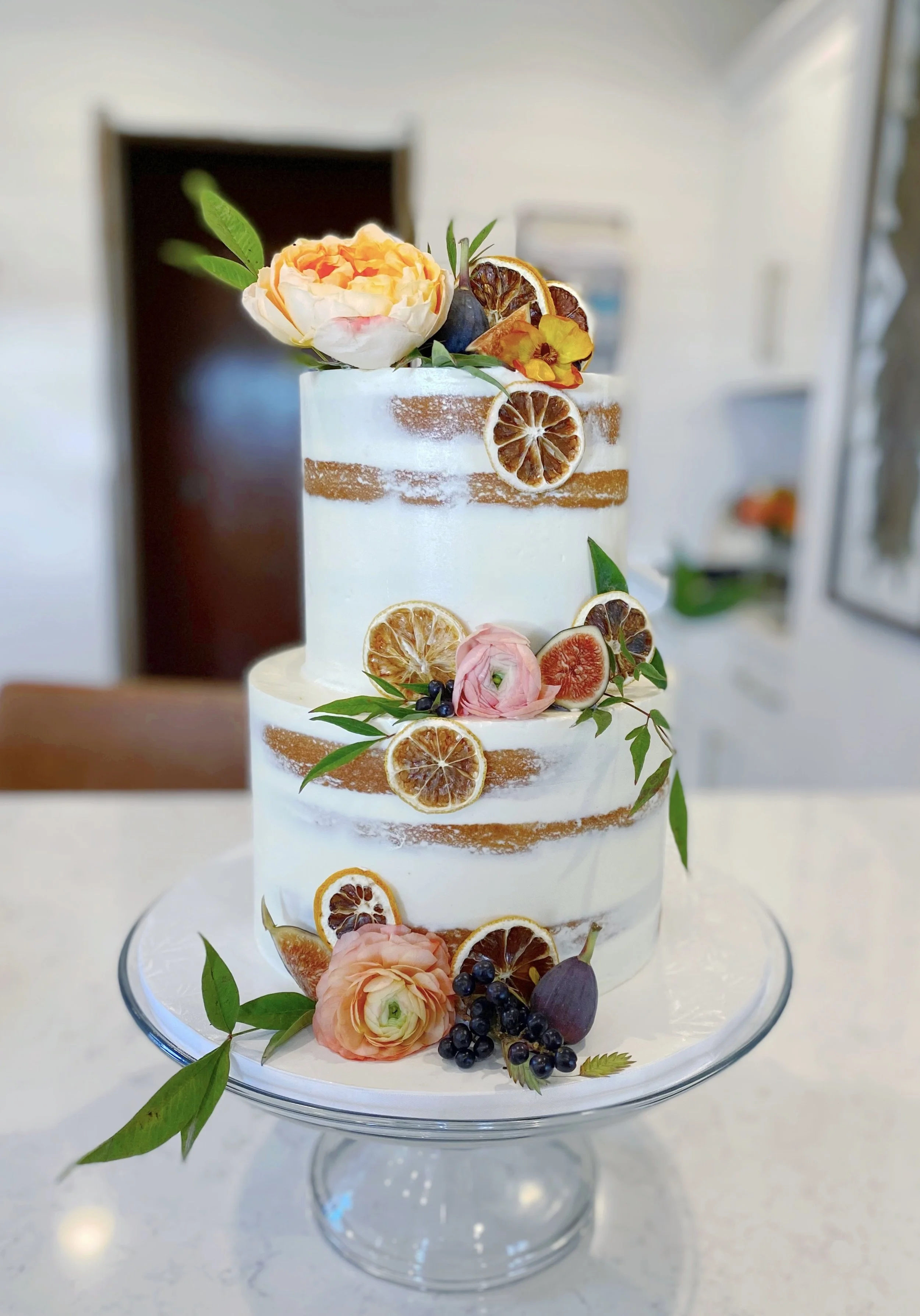 Two-tiered white naked cake decorated with dried citrus slices, fresh flowers, and green leaves on a glass cake stand.
