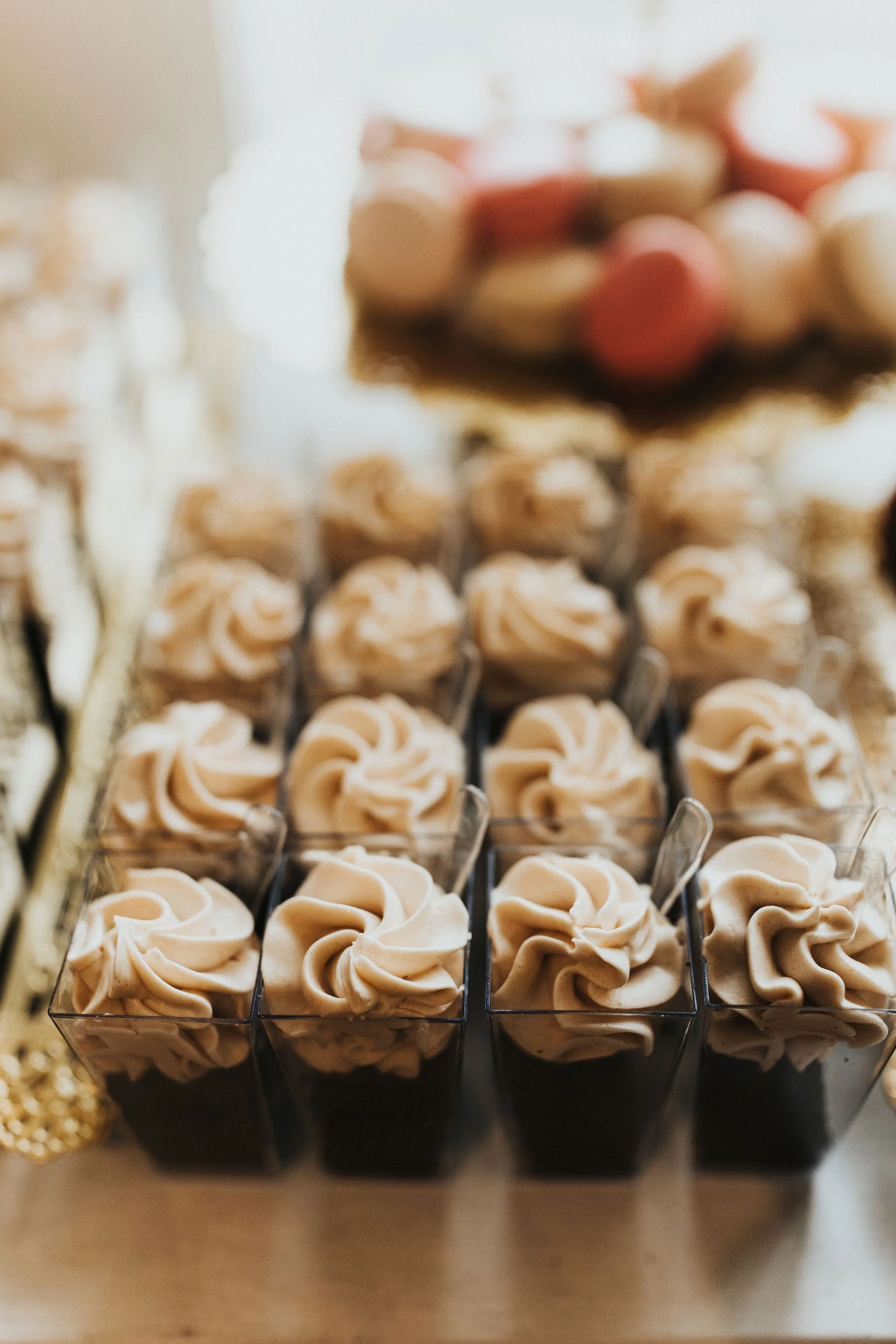 A row of five clear cups filled with a dark beverage topped with swirls of tan whipped cream or mousse, with a blurred bowl of round candies or cookies in the background.