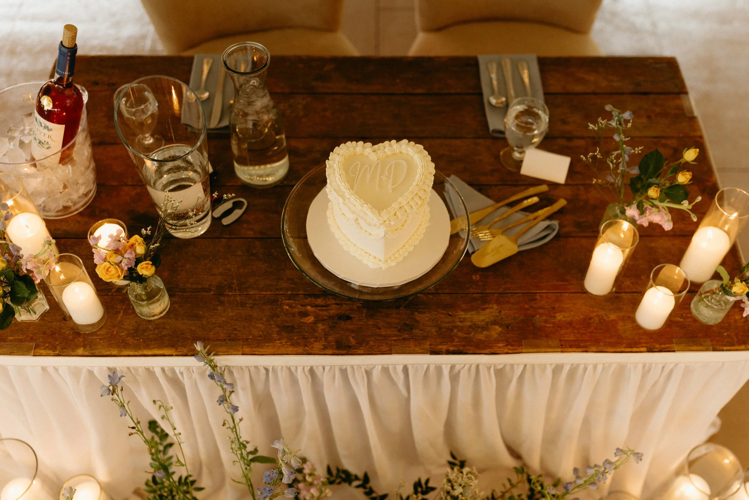 Wedding reception table with a heart-shaped cake, surrounded by lit candles, floral arrangements, and glasses of water.