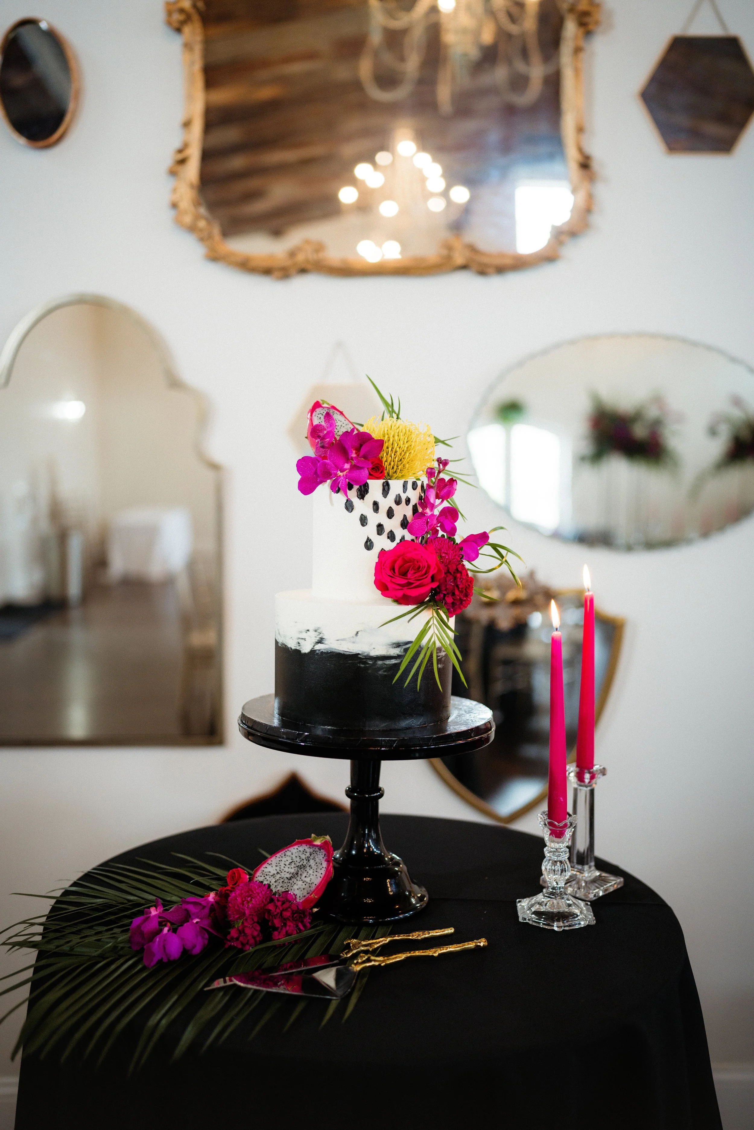 A black and white modern cake decorated with vibrant pink and yellow flowers, set on a black table with pink candles, tropical leaves, and a dragon fruit. In the background, three mirrors and a wooden ceiling are visible.