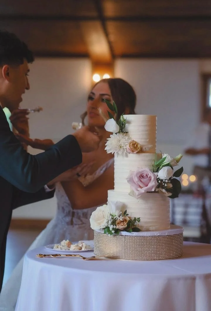 A bride and groom at their wedding cake cutting ceremony with a three-tiered white cake decorated with pink and white roses and other flowers, placed on a round table with a white tablecloth.