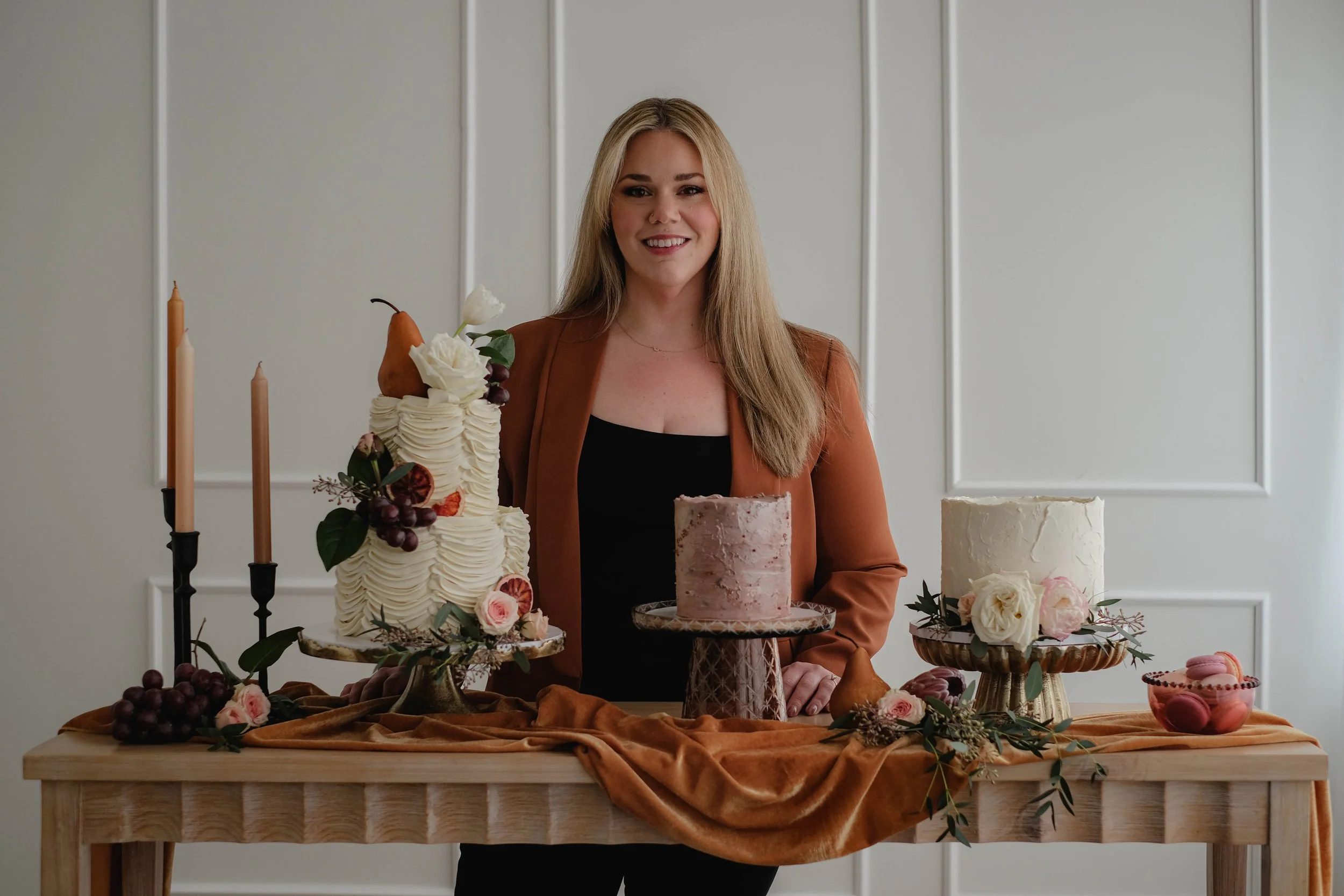 Woman standing behind a table with three decorated cakes, flowers, candles, and grapes, in a room with white paneled walls.