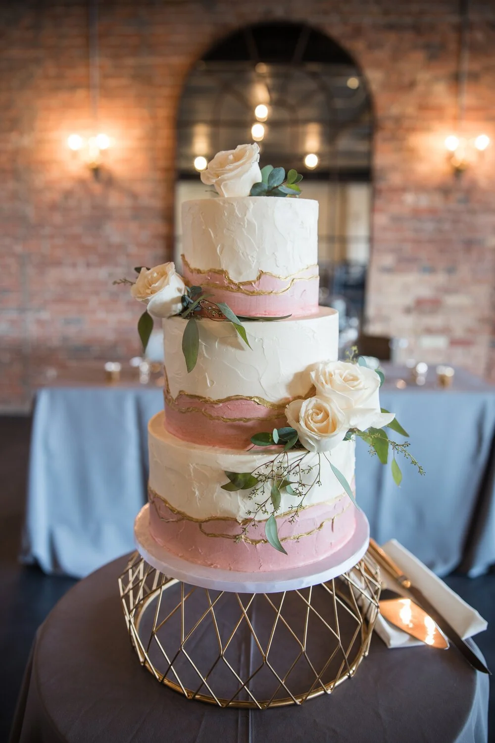 A three-tiered wedding cake decorated with white and pink frosting, gold accents, white roses, and greenery, placed on a decorative gold stand.