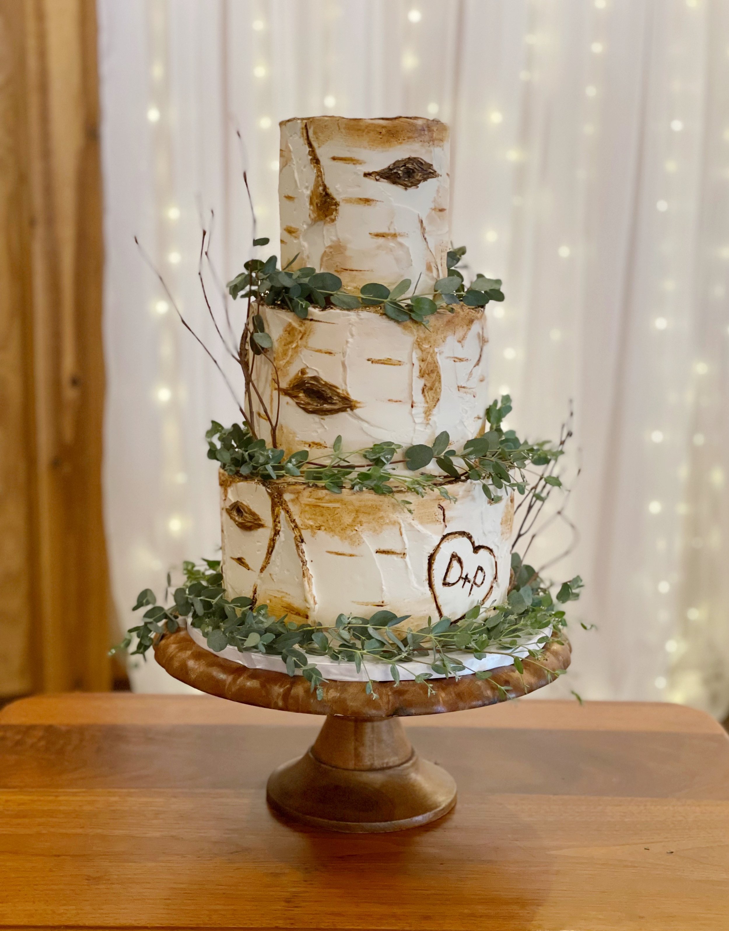 Three-tier wedding cake decorated with birch bark design, greenery, and a heart with initials 'D+O', on a wooden cake stand.