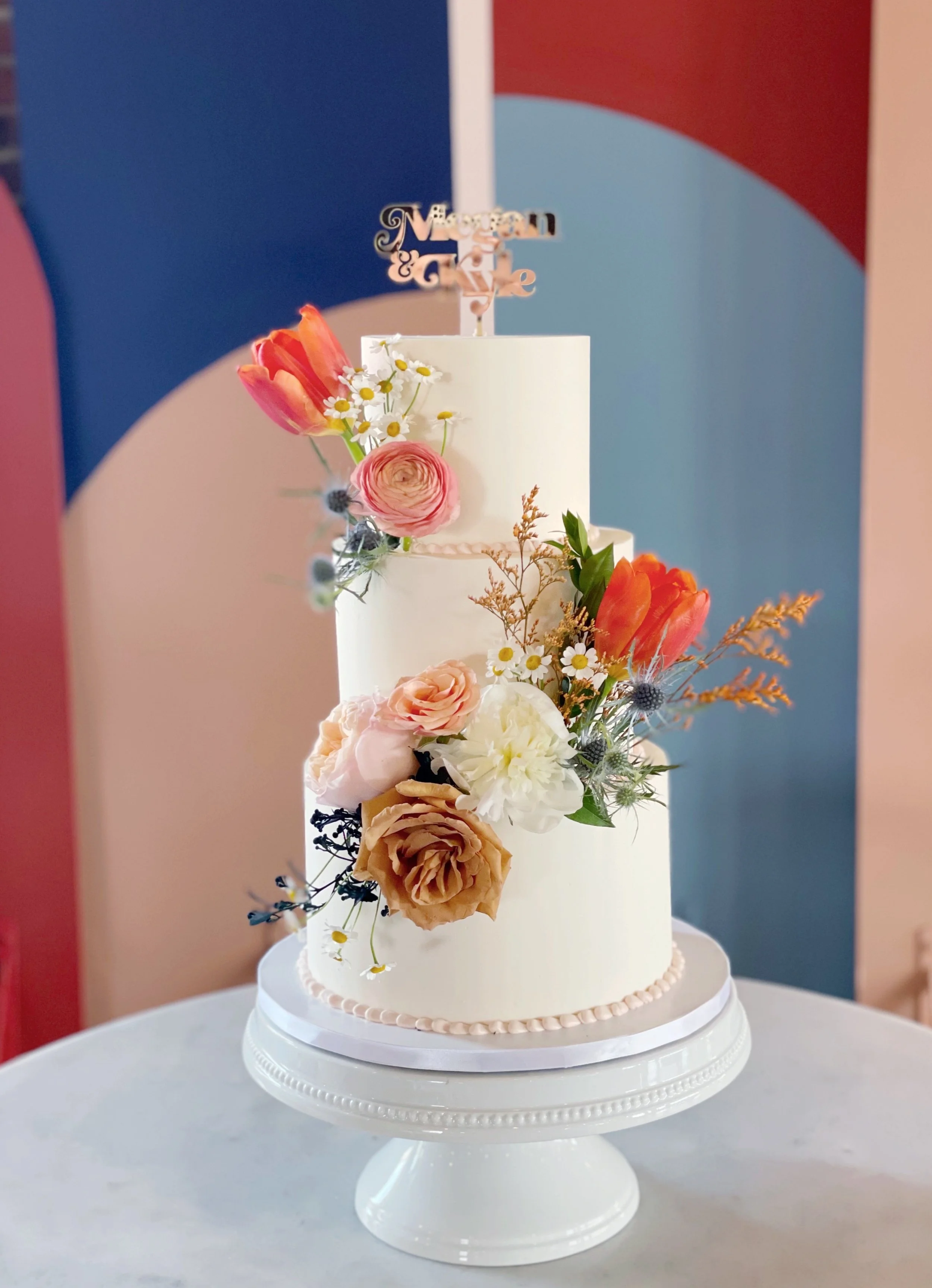 A two-tiered white wedding cake decorated with fresh flowers including pink and orange tulips, white roses, ranunculus, and small daisies, with a wooden cake topper that says 'Megan & Kyle', displayed on a white cake stand.
